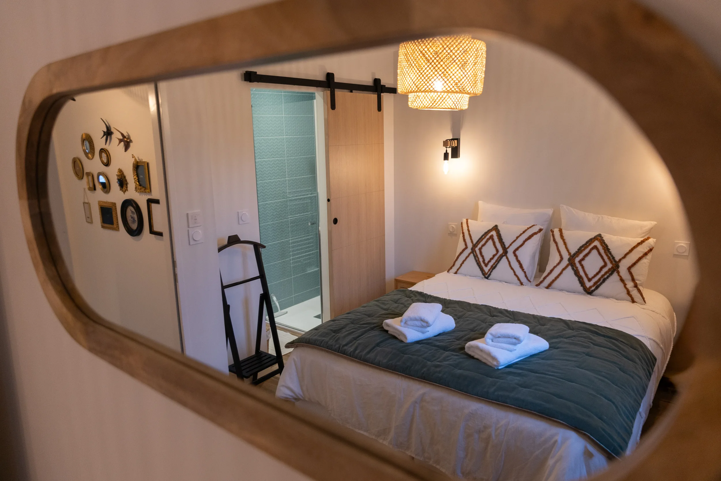 Une chambre moderne vue à travers un miroir, avec un lit fait, quatre oreillers décoratifs, deux serviettes blanches pliées sur le lit, et une porte en bois menant à une salle de bain avec murs en carrelage vert.