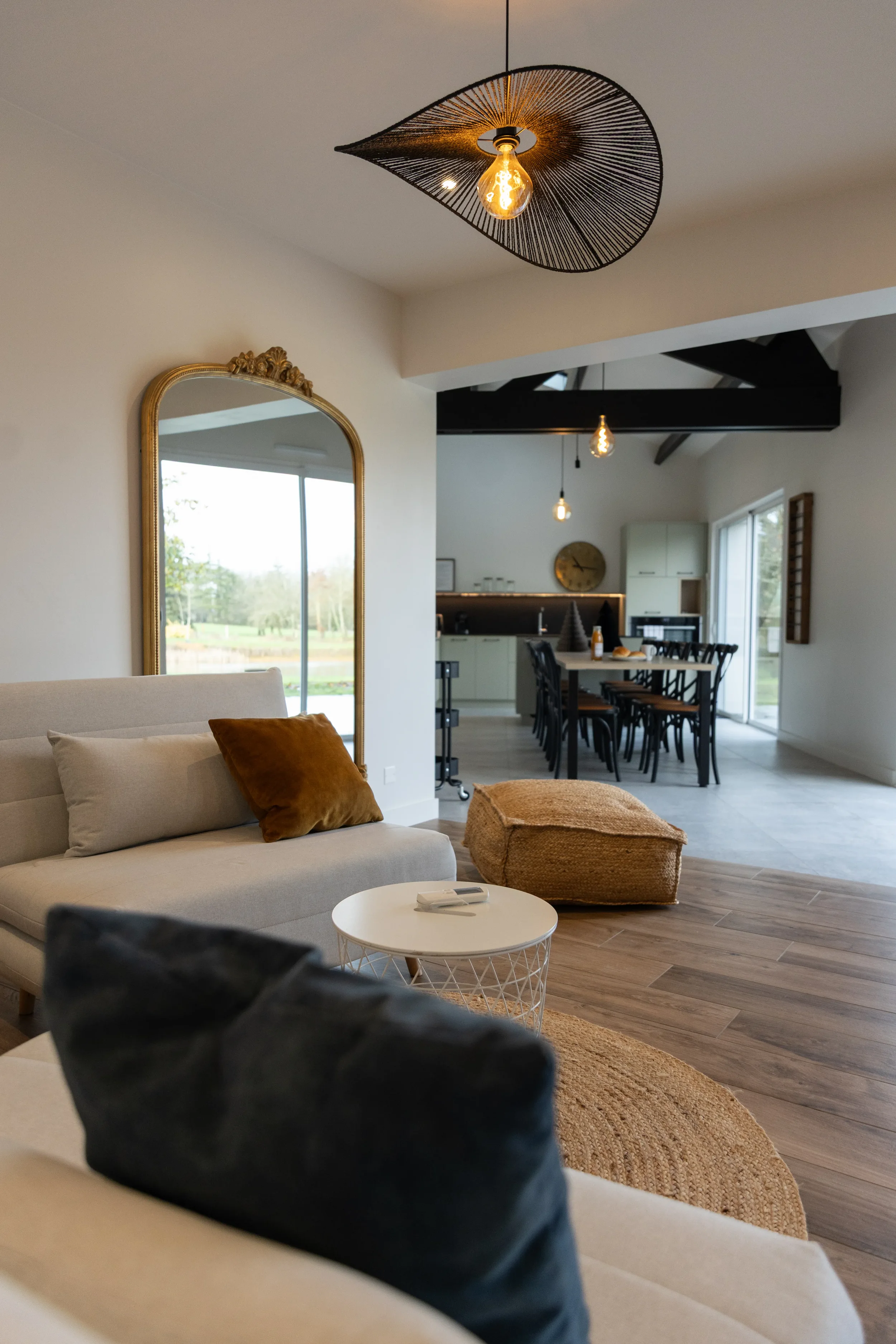 Salon moderne avec canapé beige, coussin marron, miroir ancien à cadre doré, table d'appoint ronde blanche, pouf en jute, lumière suspendue décorative. Espace ouvert avec vue sur la cuisine et la salle à manger, grande fenêtre, sol en bois et carrelage.