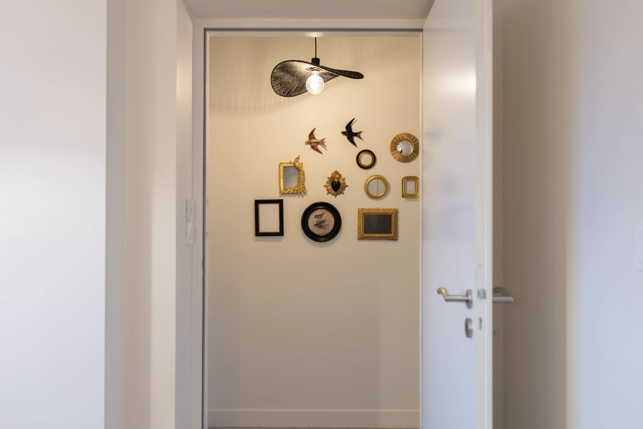 Mur blanc avec un mur de cadres et décorations murales, y compris des oiseaux en métal, un miroir rond avec une étoile de mer, et un cadre noir contenant deux oiseaux dessinés, éclairé par une suspension avec une ampoule.