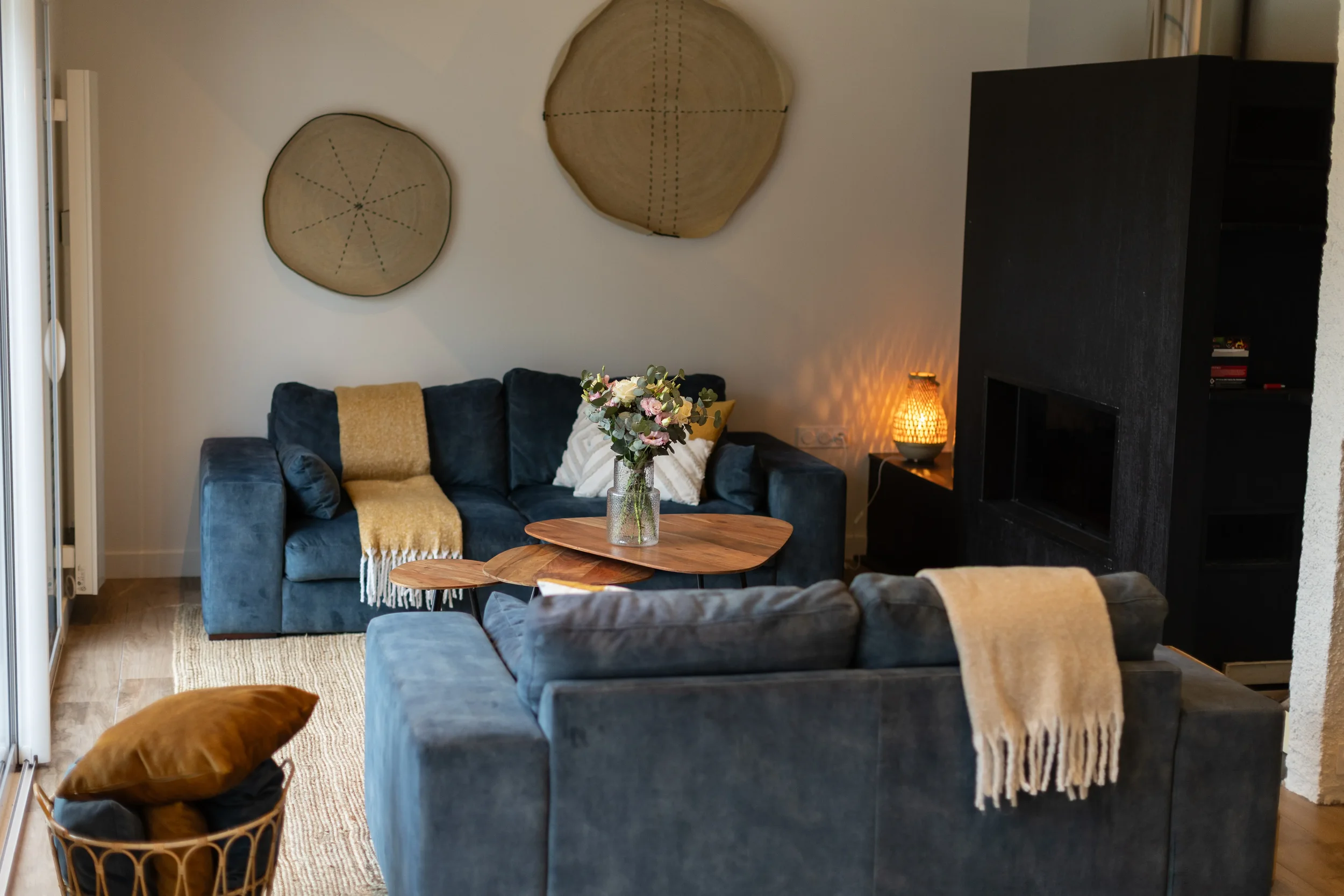 Salon avec deux canapés en velours bleu, table en bois avec bouquet de fleurs, mur blanc avec deux plaques en bois décoratives, lampe diffuse à côté de cheminée noire