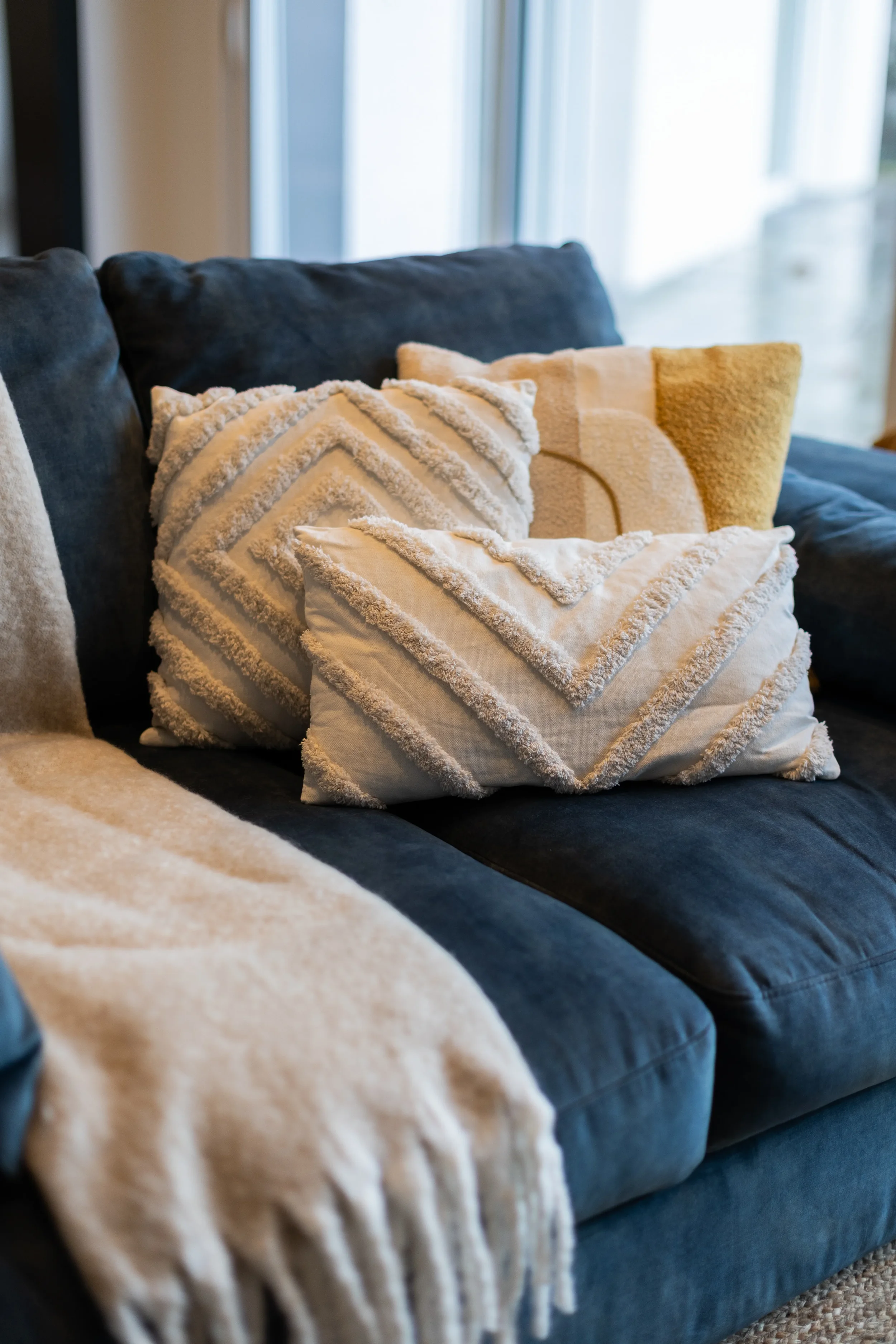 Coussins décoratifs en tissu beige et blanc avec motifs géométriques et textures épaisses, posés sur un canapé en tissu bleu foncé près d'une fenêtre