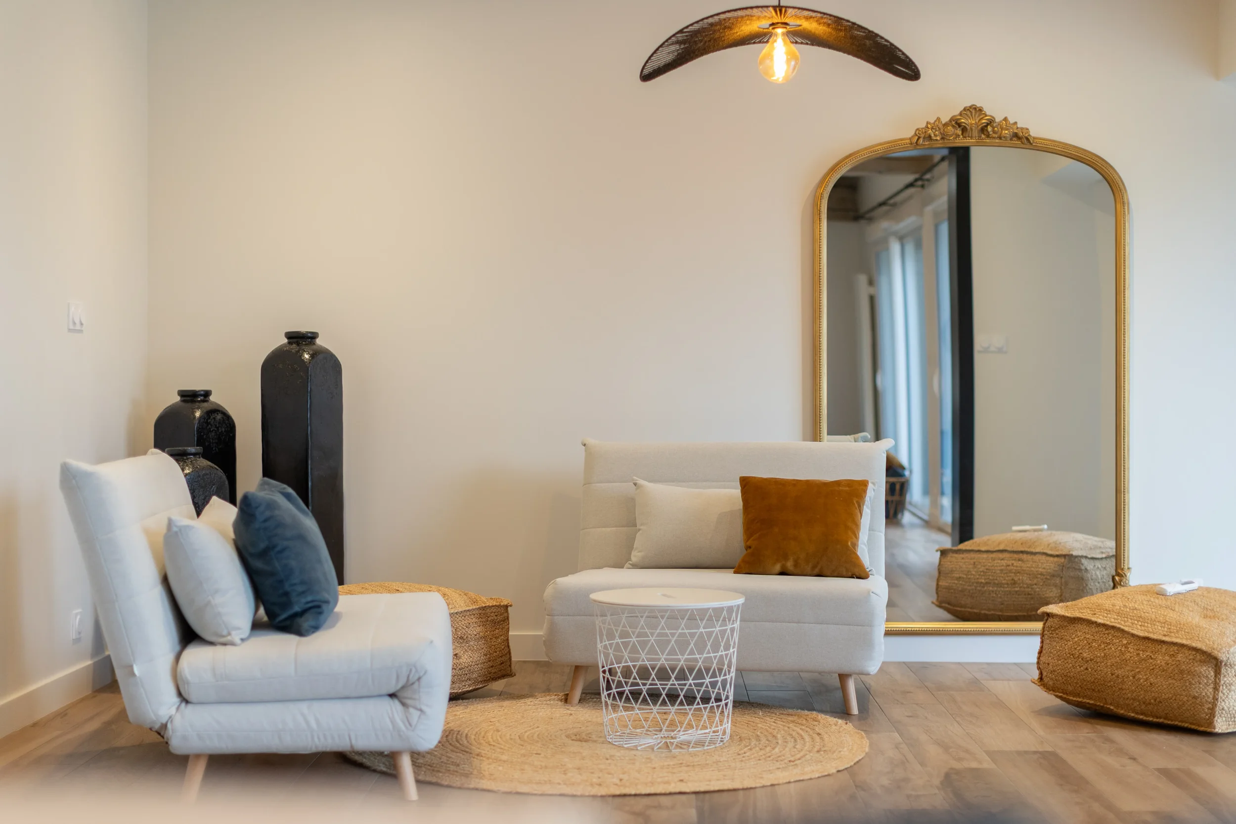 Salon moderne avec deux sofas, une table ronde en métal blanc, un grand miroir doré et des vases noirs, dans un décor épuré