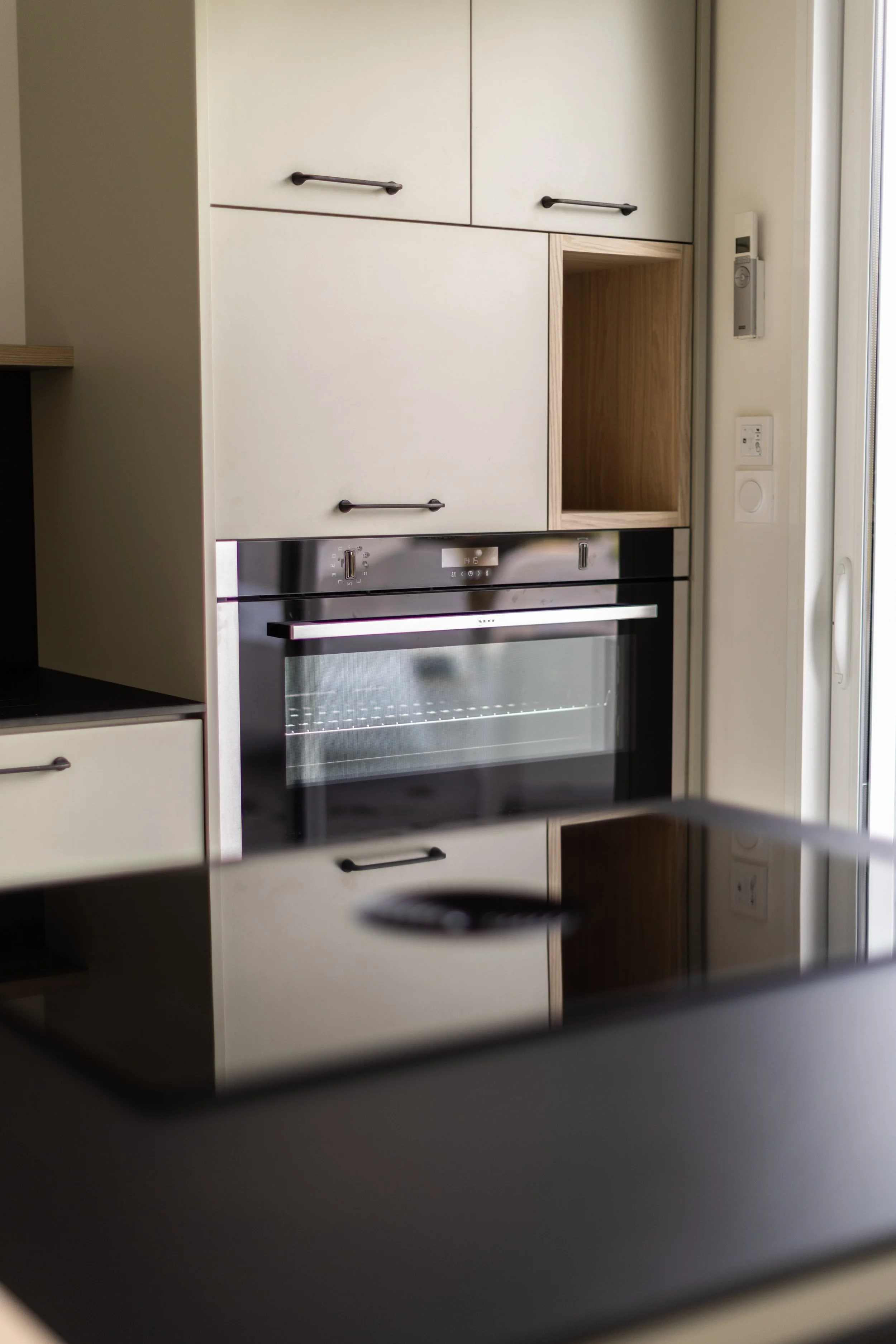 Vue d'une cuisine moderne avec un four électrique encastré et des armoires en bois clair ou beige. La photo est prise depuis un angle, avec des éléments de la pièce visibles en reflet dans la porte du four.