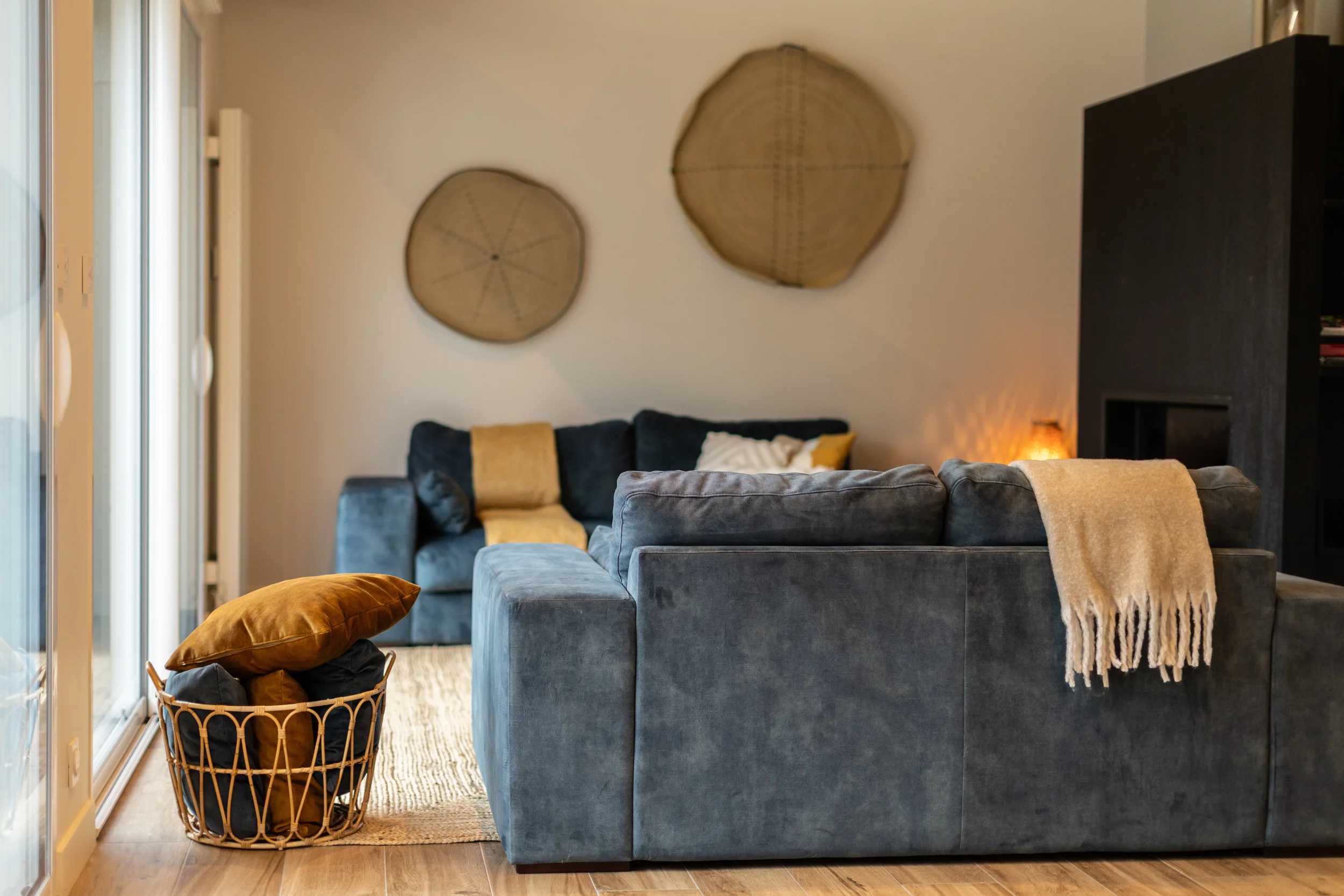 Salon avec canapé en velours gris, coussins beiges et jaunes, tapis beige, décor mural en bois, panier à linge avec coussins, éclairage chaleureux.