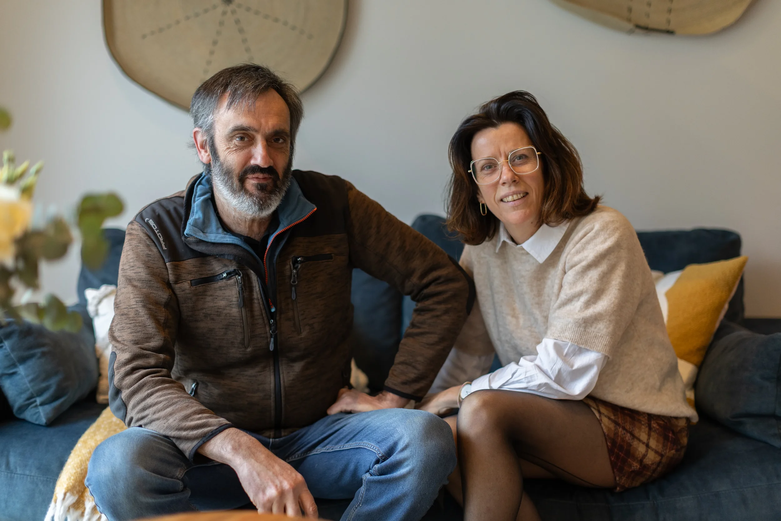Un homme et une femme assis sur un canapé dans un salon, posant pour une photo. L'homme porte une veste brune et la femme porte un pull beige avec une chemise blanche. Ils ont l'air détendus et souriants.