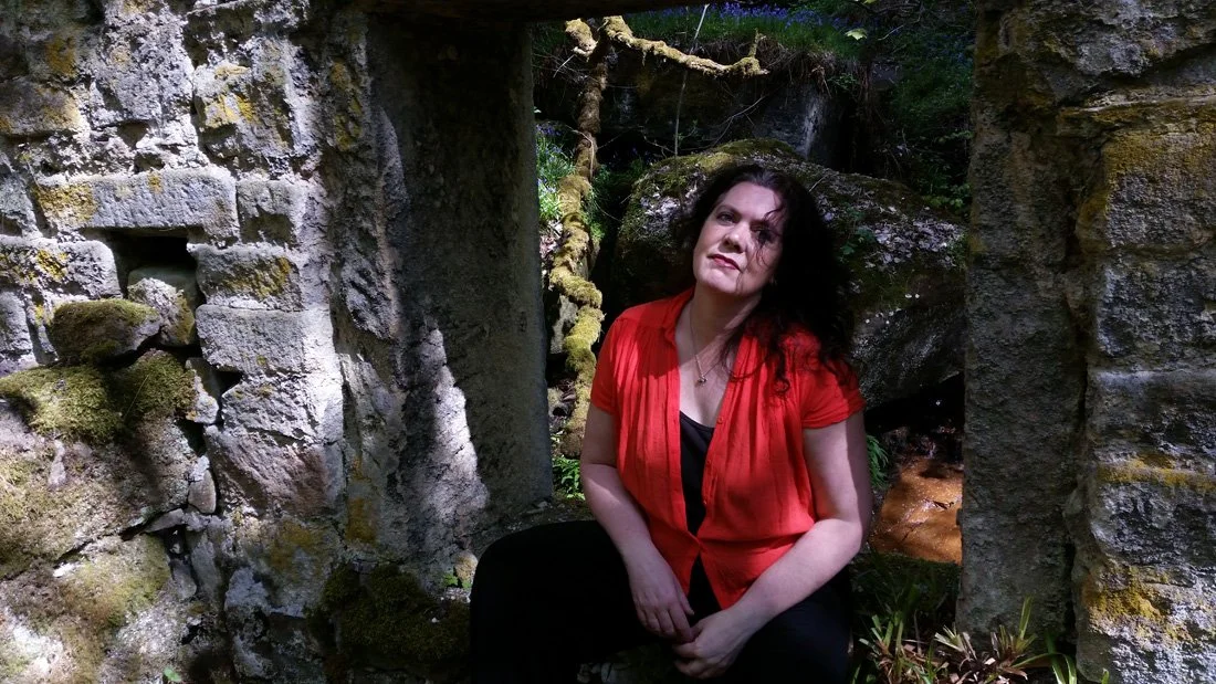 A woman with curly dark hair in a red and black outfit sitting between stone ruins in a forest with moss-covered rocks and green foliage in the background.
