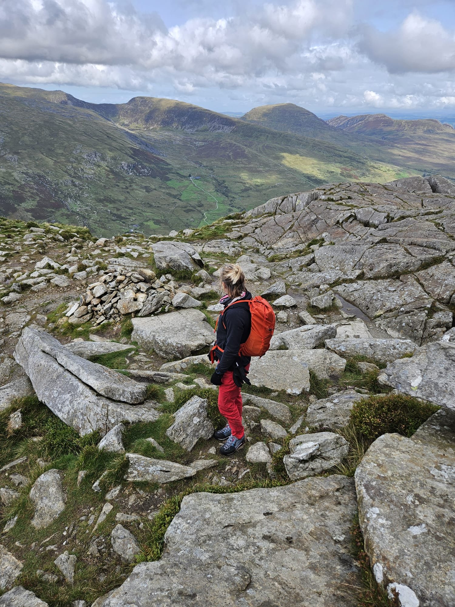 Hiker with red backpack and red pants walking on rocky mountain trail with green valley and hills in the background under cloudy sky.