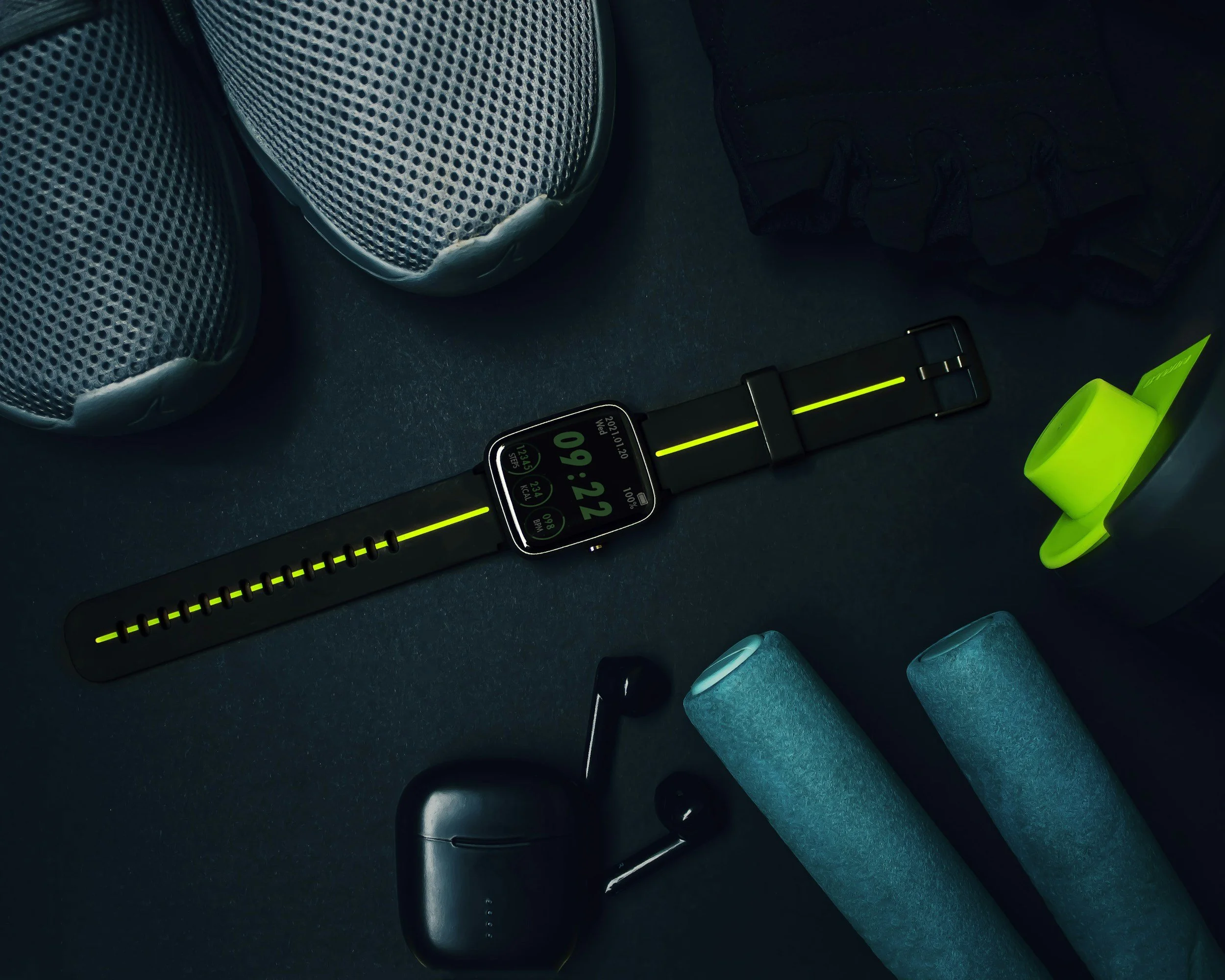 Close-up of a smartwatch displaying the time 09:20, surrounded by fitness equipment including black wireless earbuds, a black case, two foam rollers, a pair of athletic shoes, and a neon green tape dispenser on a dark surface.