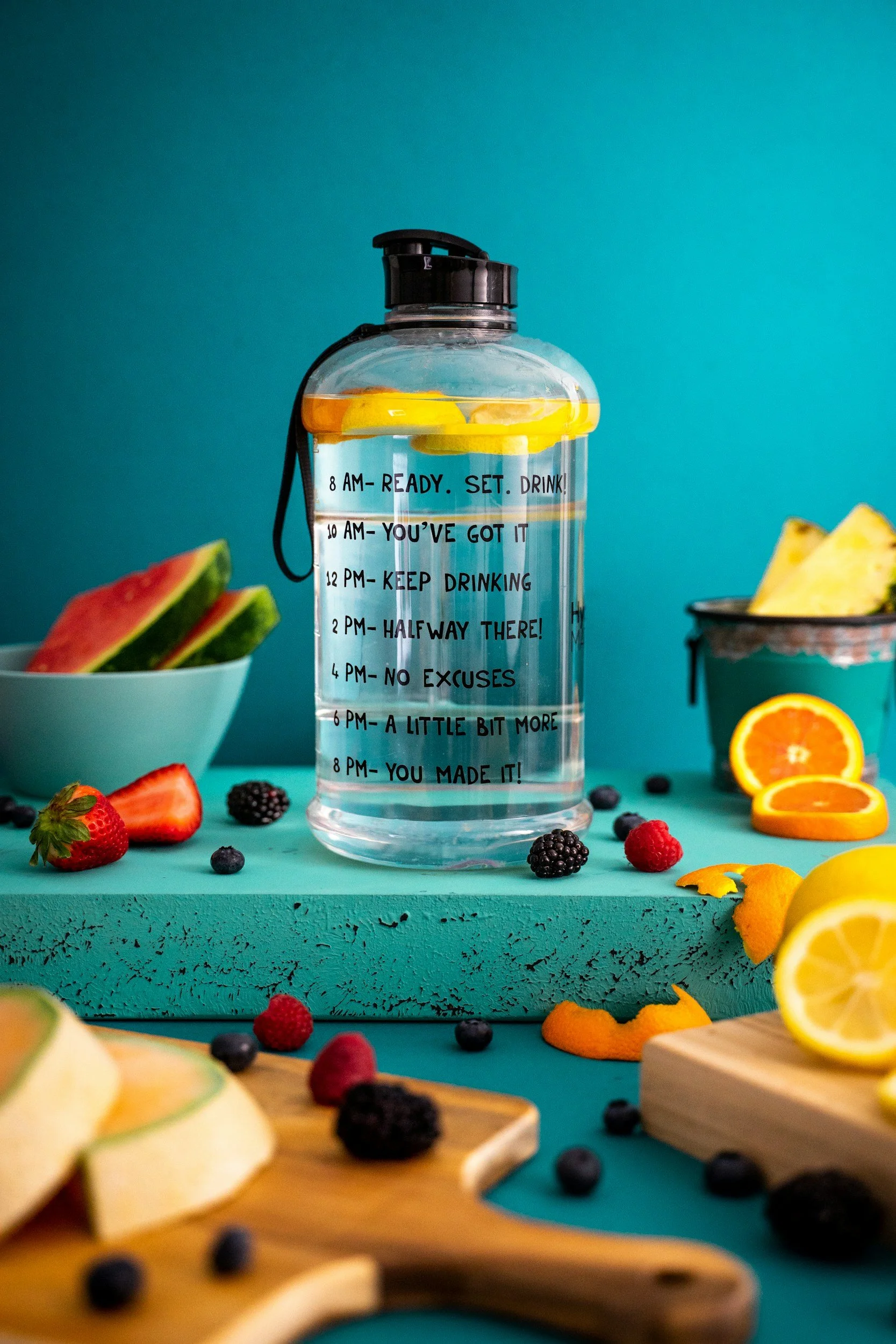 A clear water bottle with a black cap, filled with water and lemon slices, standing on a teal surface with various fruits around it. The bottle has motivational time-based messages written on it, from 8 AM to 8 PM, encouraging drinking water throughout the day.