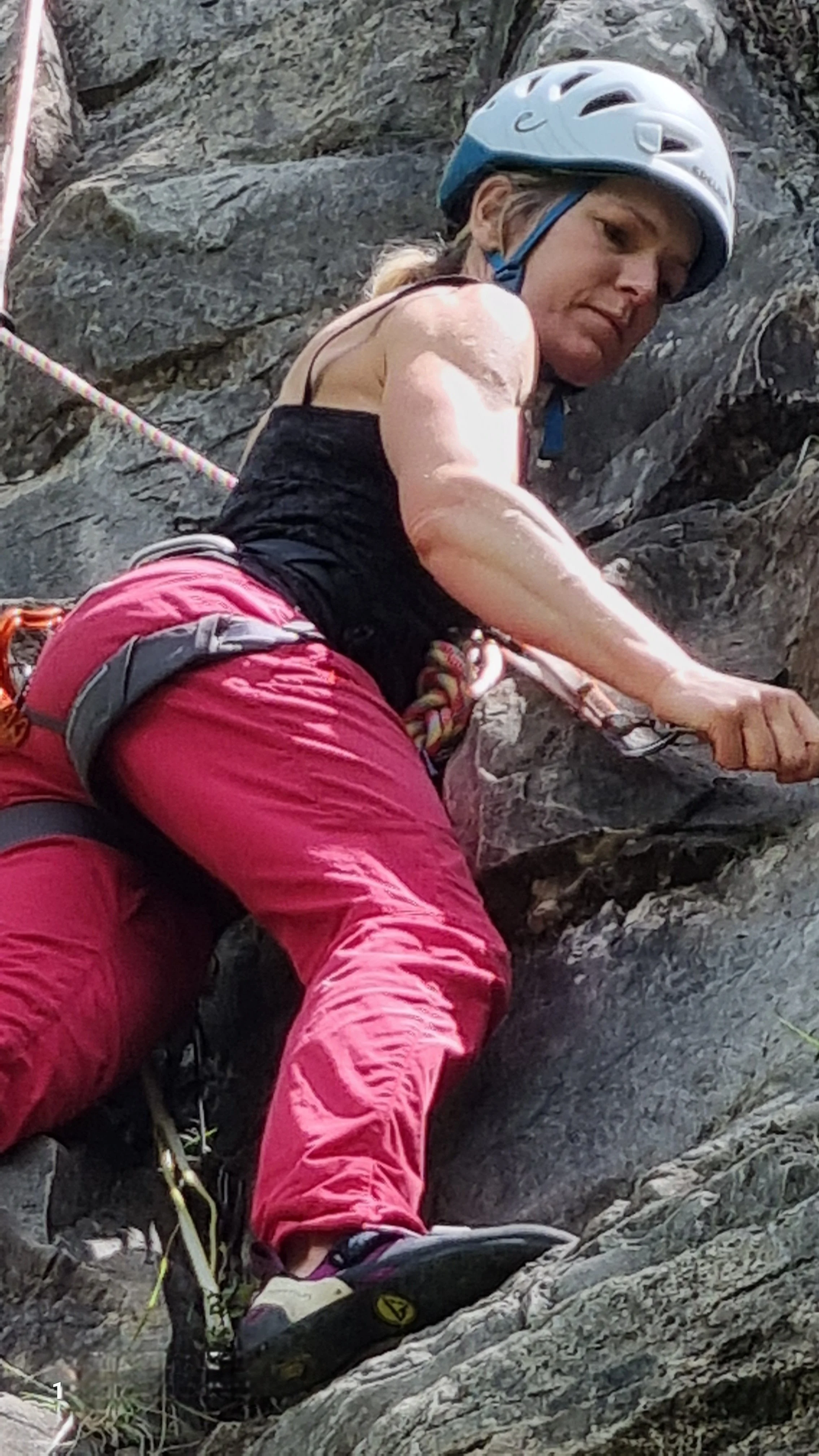 A woman wearing a white climbing helmet, black tank top, and red pants is rock climbing on a rugged, steep rock face, utilizing her hands and feet to ascend.