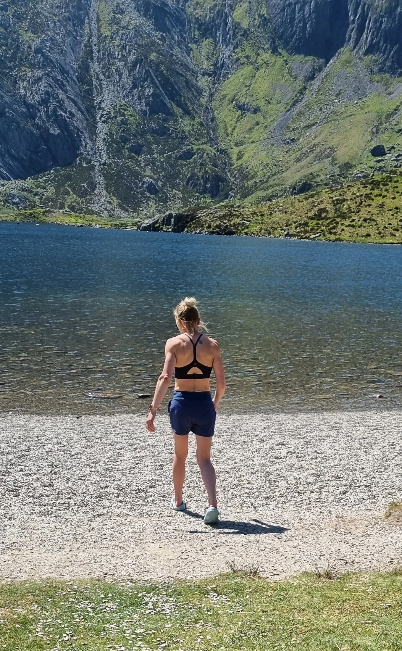 A woman in a black sports bra and blue shorts walking towards a lake with steep, green mountains in the background.