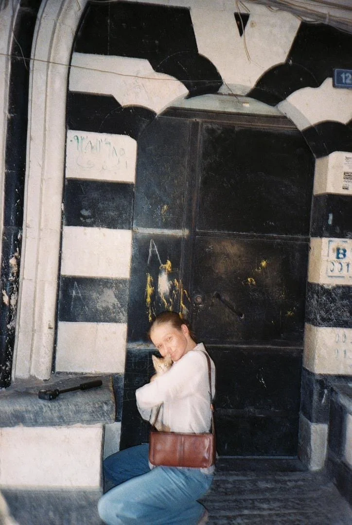 A young woman sitting on a bench hugging a small dog in front of a black and white striped door with an arched top, in an urban setting. Emilysfamous syria tour