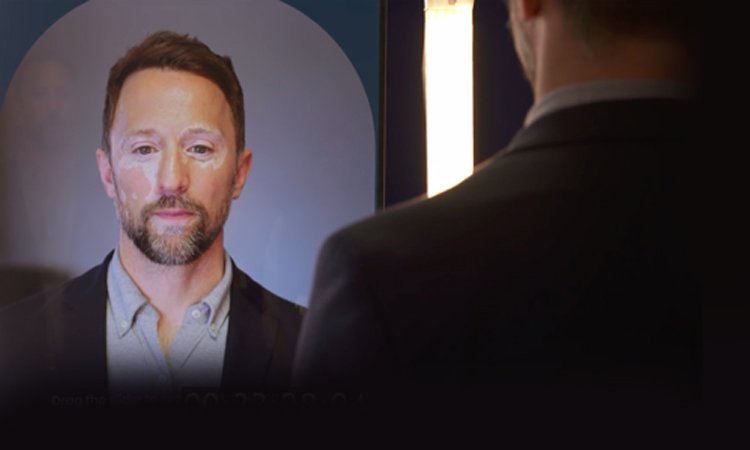 A man with a beard and short brown hair looking at a mirror or screen, with his reflection visible, in a professional setting.