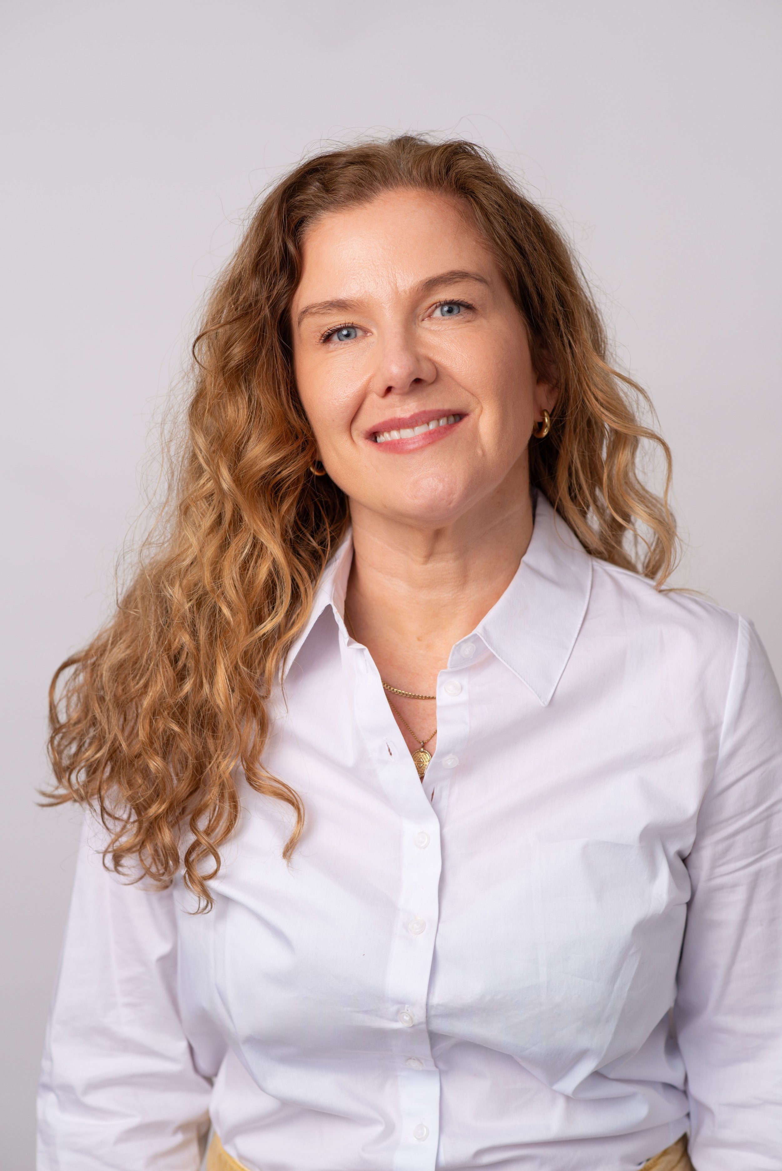 Portrait of a woman with long curly red hair wearing a white button-up shirt and gold jewelry, smiling against a plain gray background.