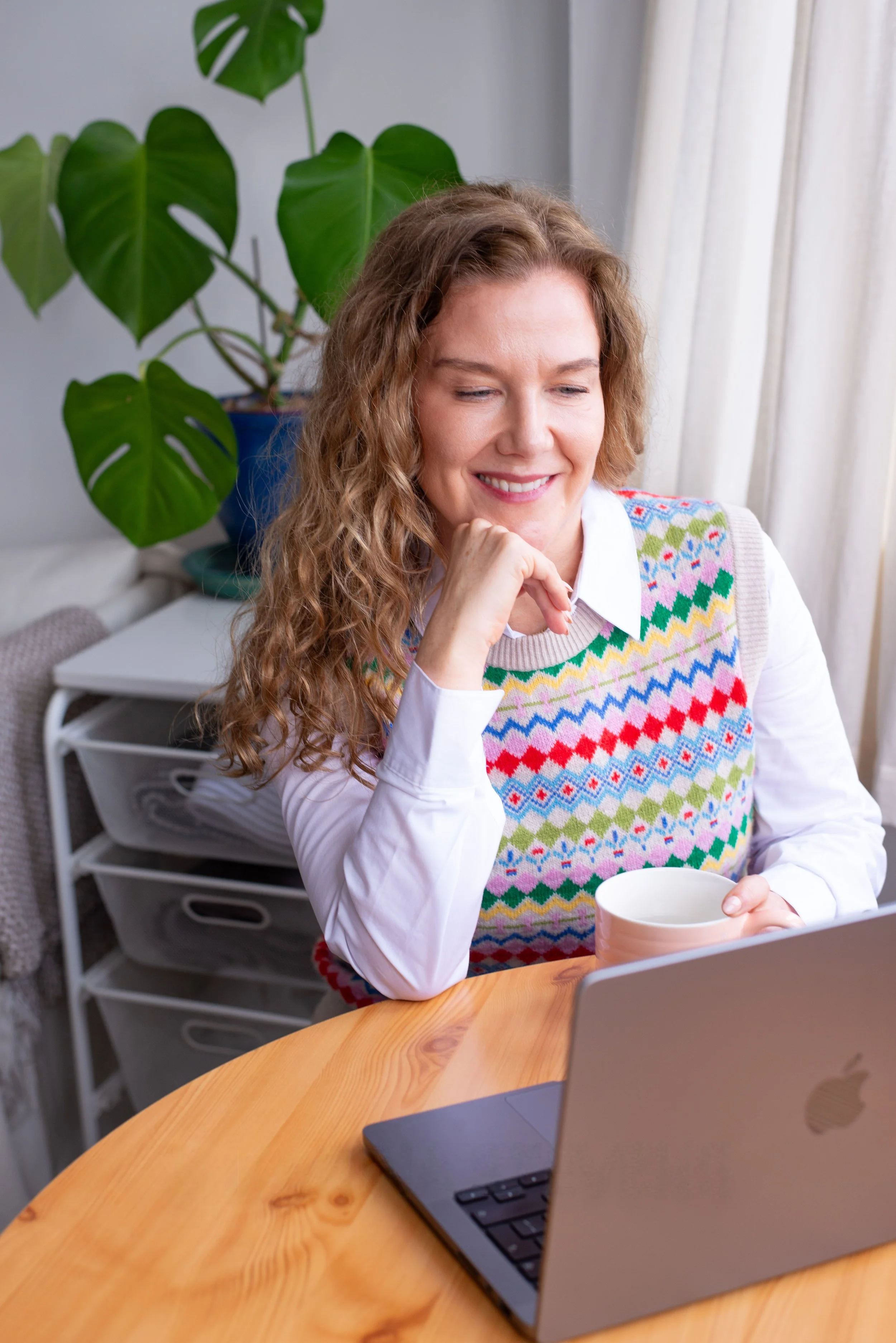 A woman with curly, light brown hair smiling and holding a pen and mug, working on her laptop at a round wooden table in a well-lit room with a large leafy plant and white curtains in the background.