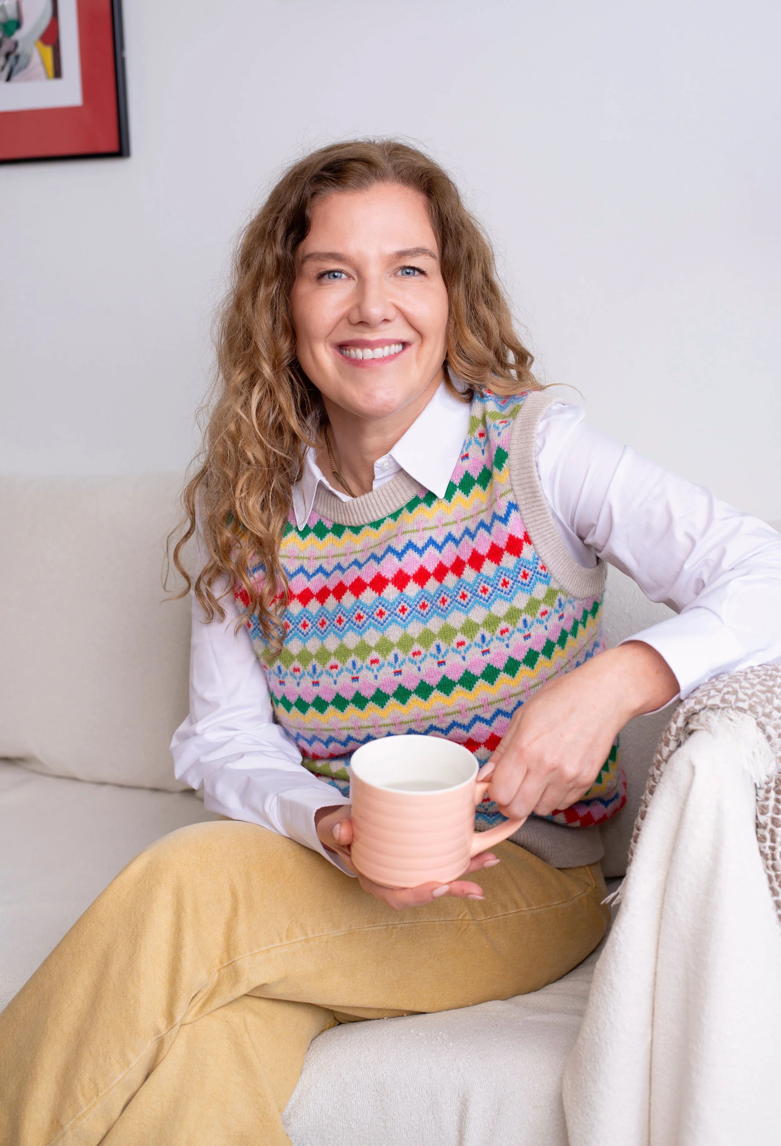 A woman with curly hair, wearing a white shirt and a colorful patterned sweater vest, sitting on a light-colored sofa, smiling, while holding a pink mug.