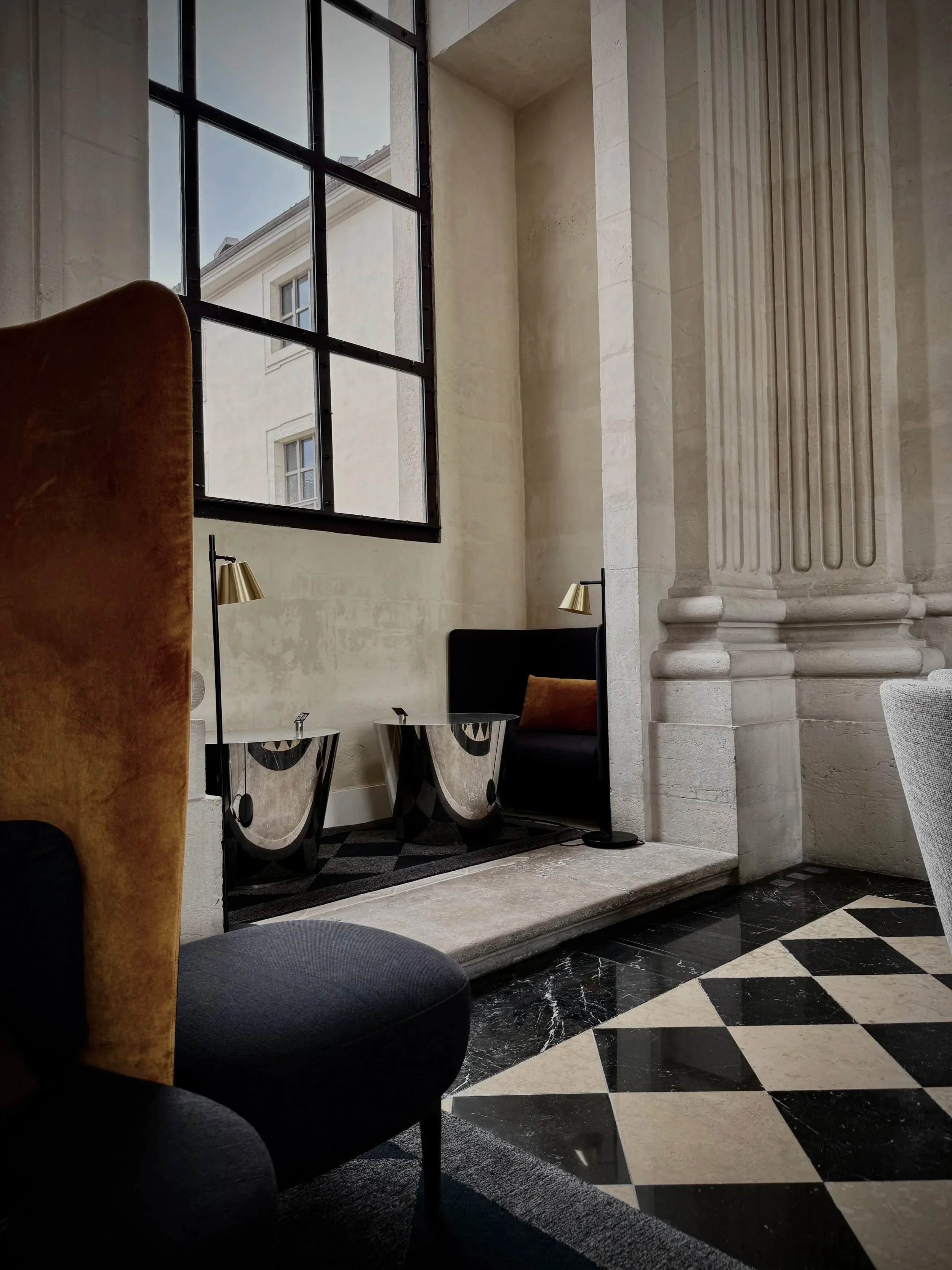 Intérieur d'un bâtiment avec un grand fenêtre en métal noir, un fauteuil en peluche marron, deux lampes modernes dorées, une table mosaïque avec miroirs, murs en pierre, sol en marbre noir et blanc avec un motif géométrique.
