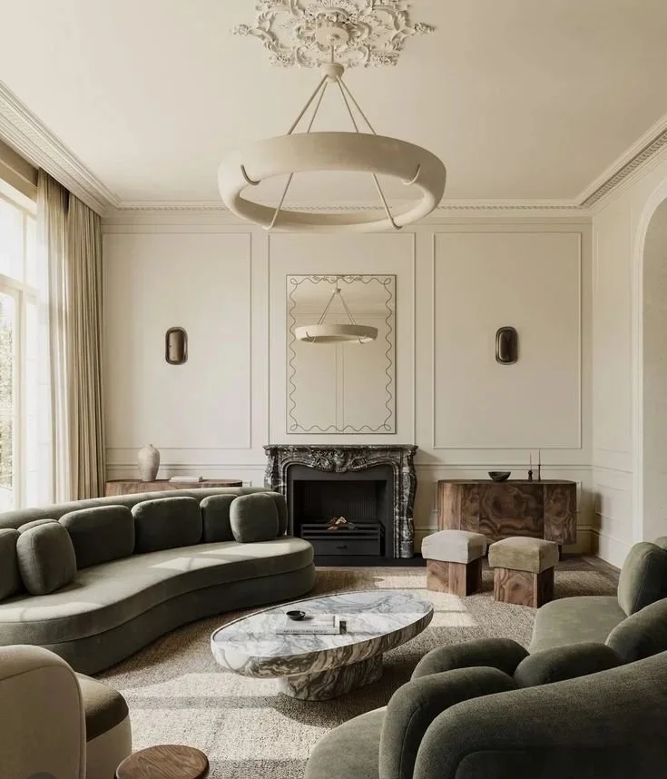 Salon élégant avec cheminée en marbre noir, grand miroir carré encadré, canapés verts en tissu, table basse en marbre, meubles en bois, grands rideaux beige, luminaires modernes, plafond orné de moulures.