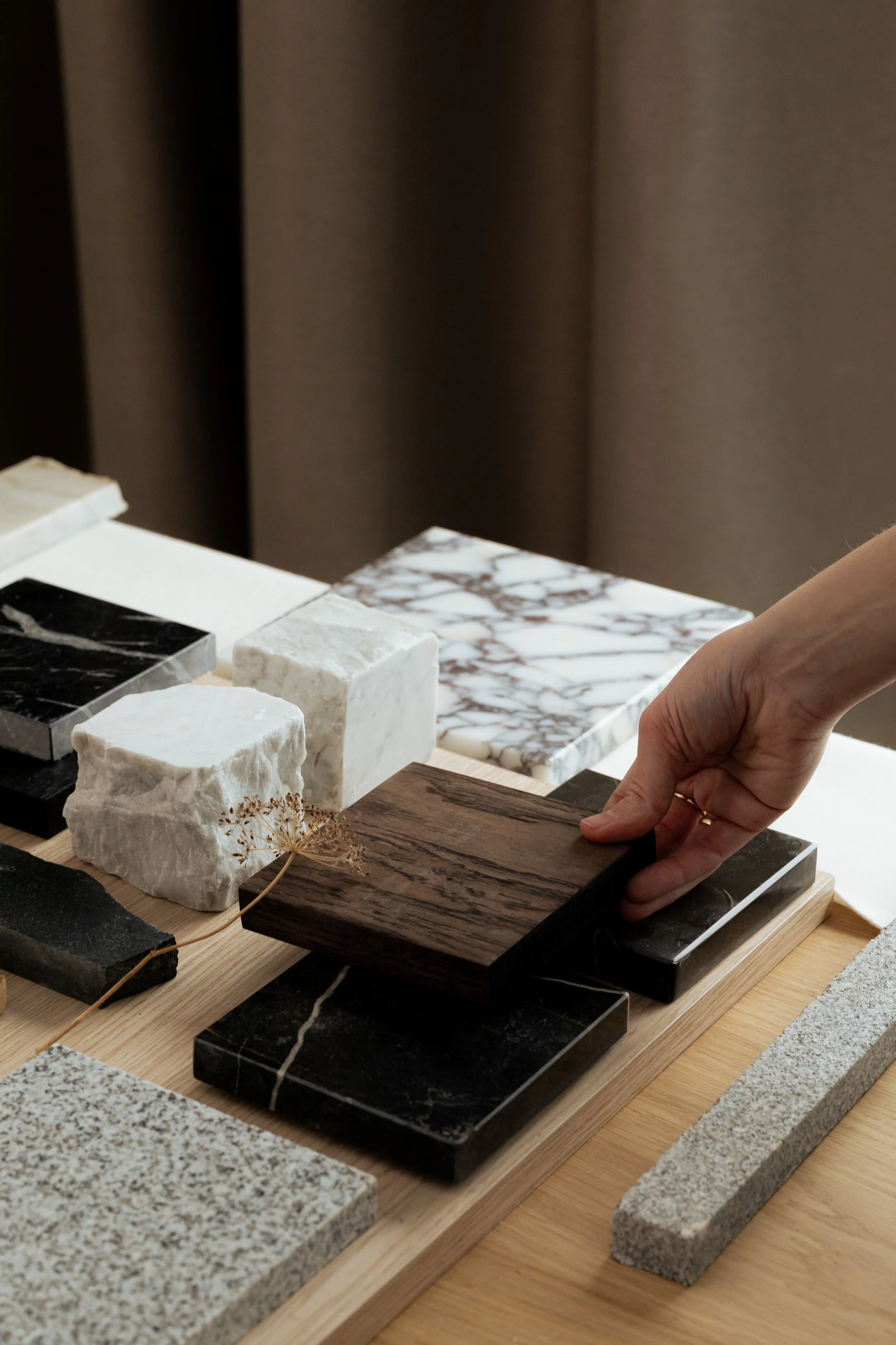 Une main touche différentes dalles de matériaux variés, posées sur une planche en bois, avec un fond de rideaux beige.
