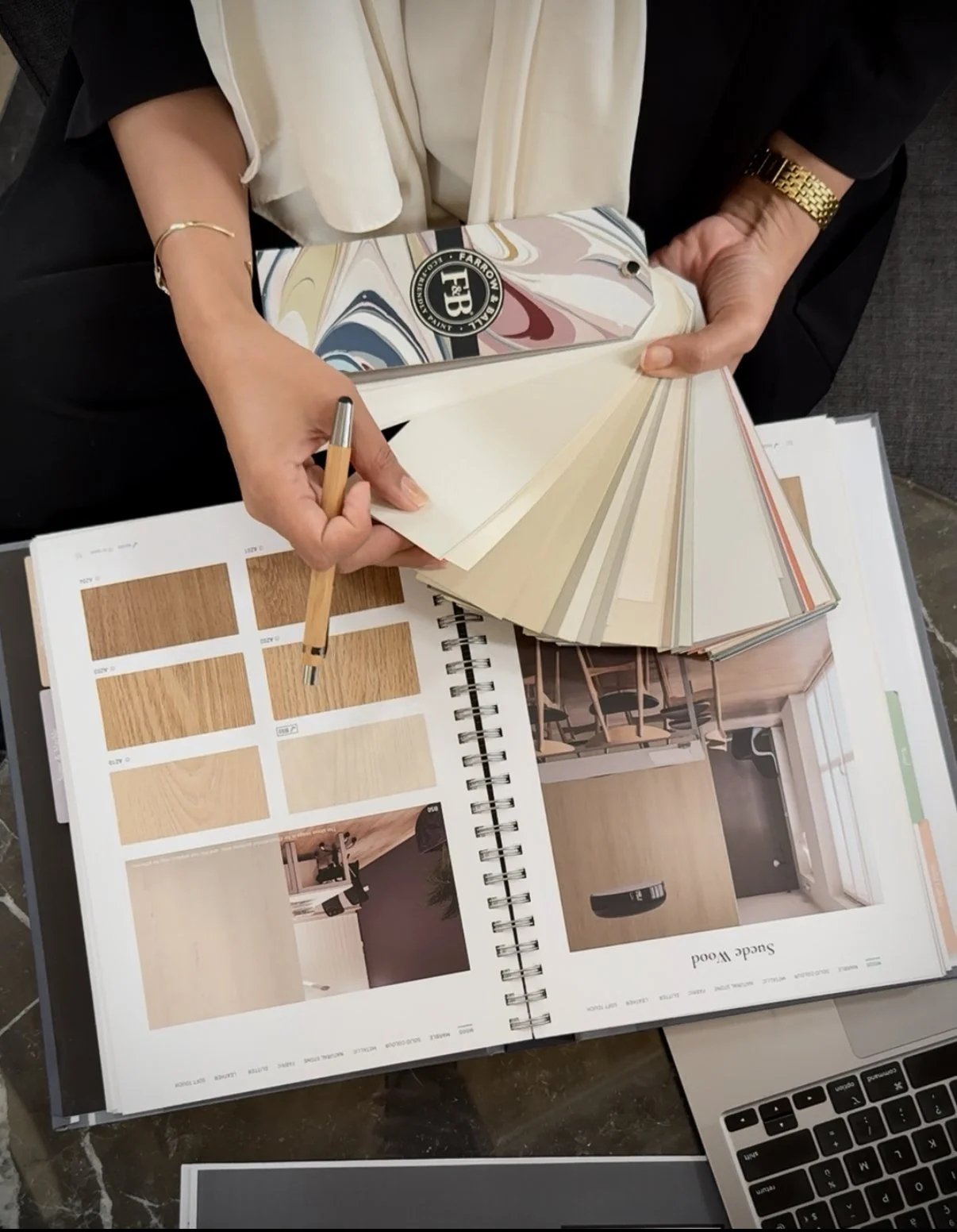 Personne tenant un échantillonnage de couleurs et un carnet de planification de décoration intérieure, avec des échantillons de bois, des photos de pièces et un ordinateur portable sur une table.
