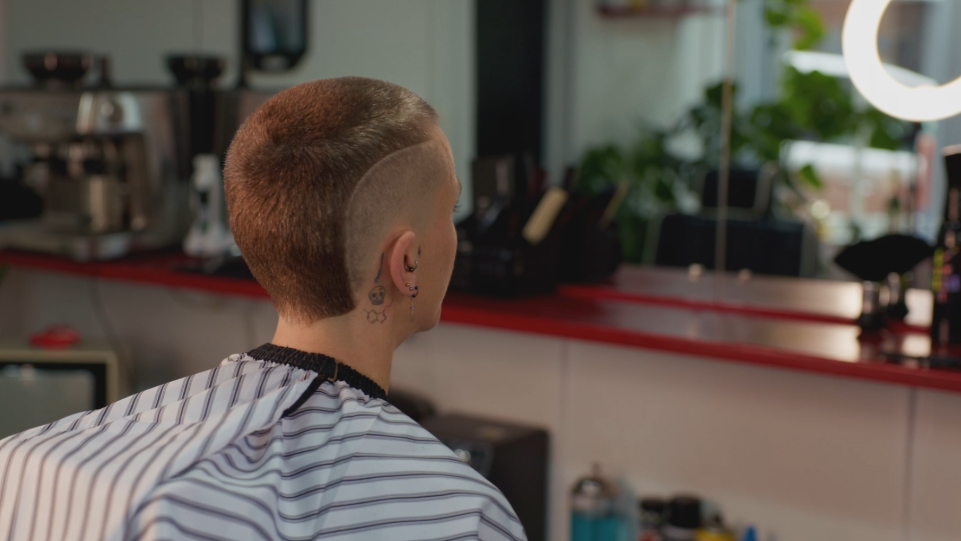 Person with a short tan hairstyle and multiple earrings sitting in a salon, facing away from the camera, with a background of salon equipment and a mirror.