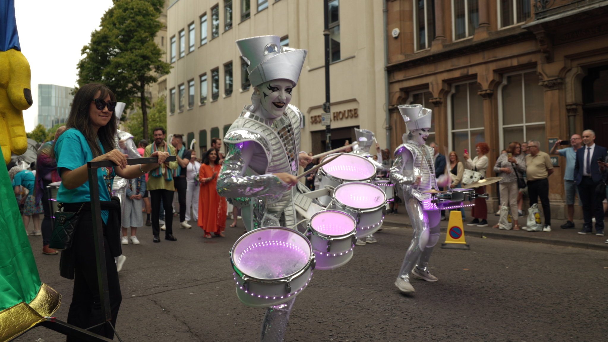 Three performers dressed as futuristic, metallic musicians with glowing drums marching in a parade, with onlookers watching on the street.