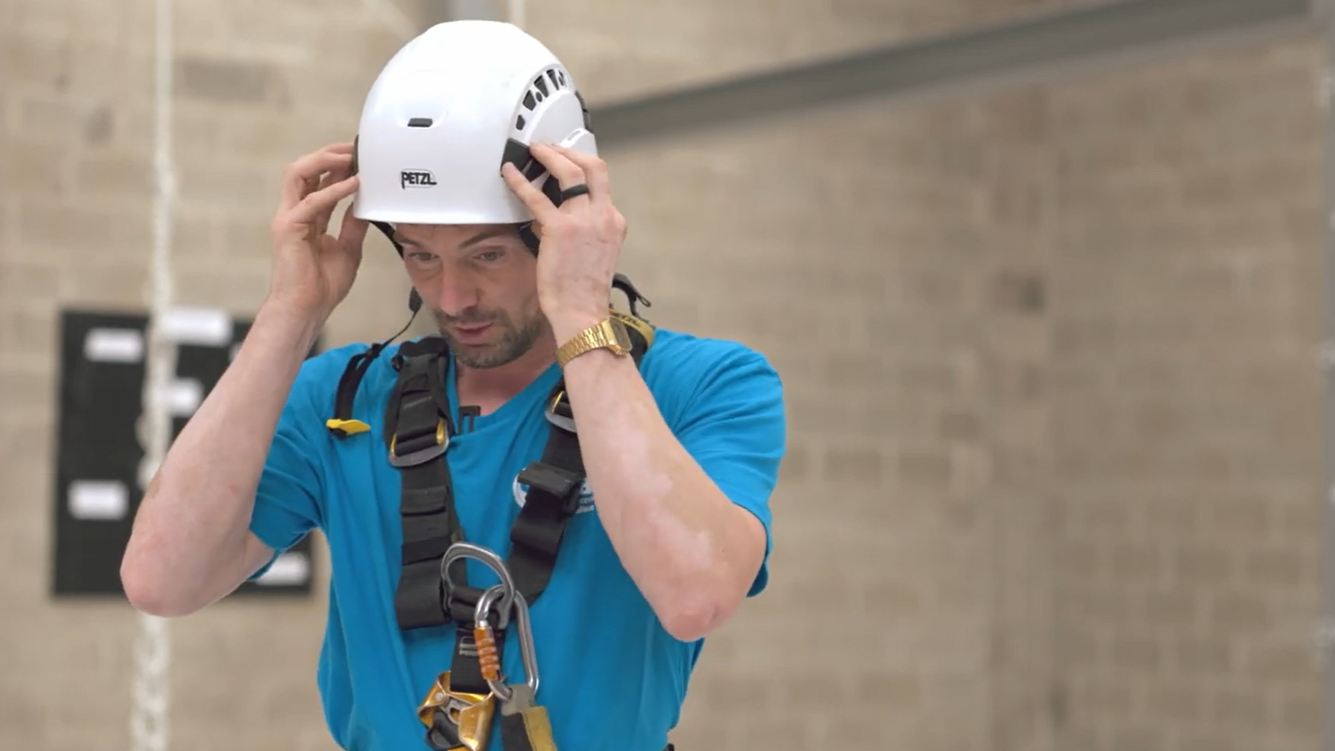 A man wearing a construction helmet and climbing harness adjusts his helmet.