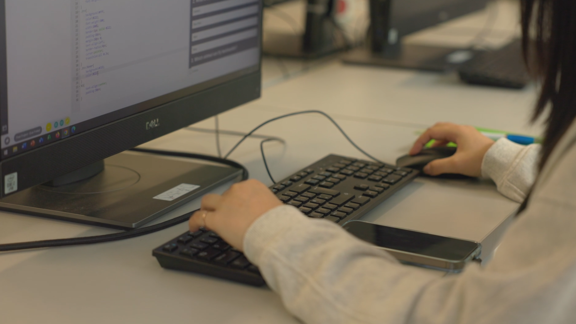 Person working at a computer with code on the screen, using a keyboard and mouse, with a smartphone placed nearby on the desk.