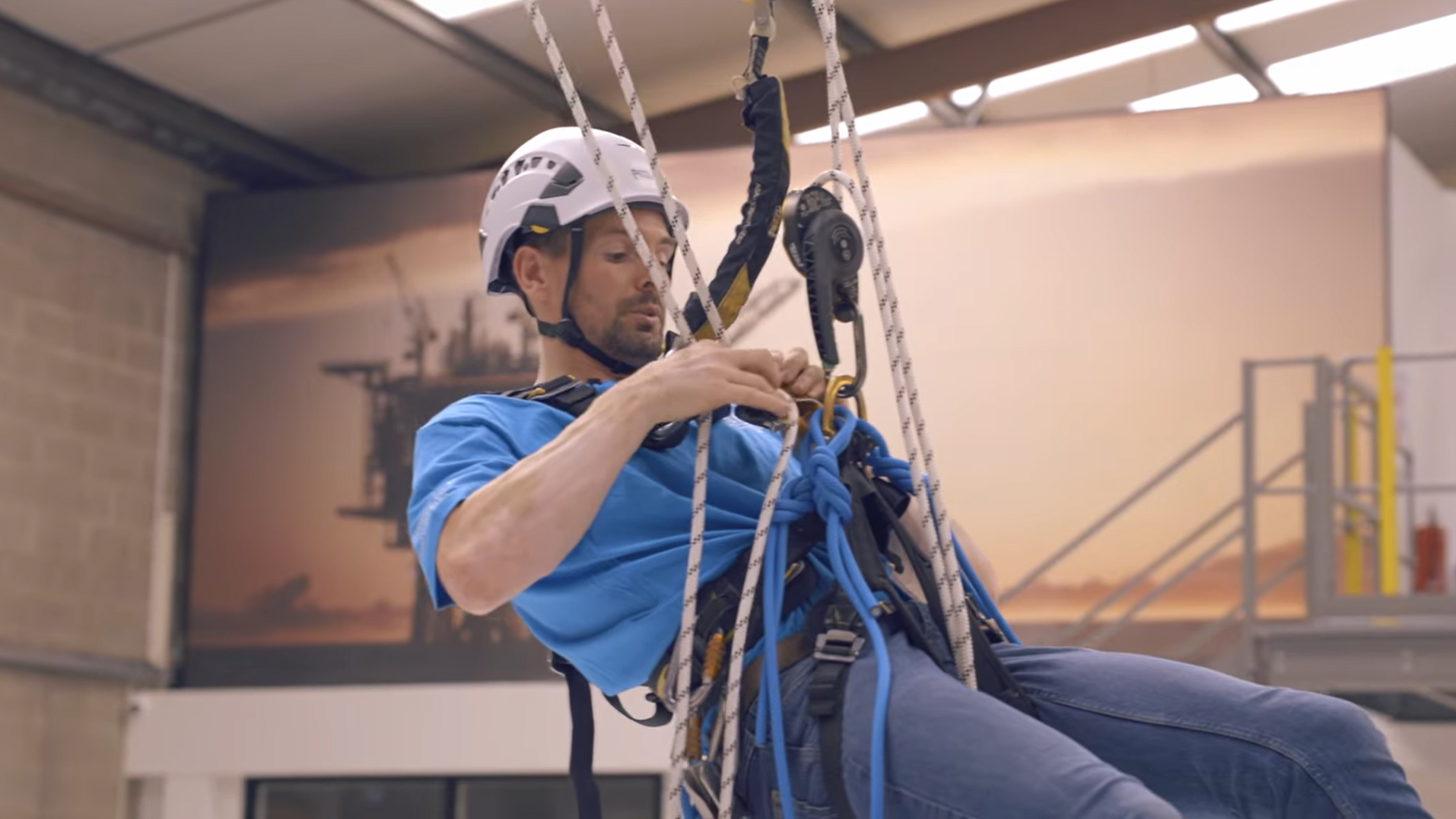A man wearing a safety helmet is adjusting climbing gear attached to ropes and harnesses in an indoor training facility.