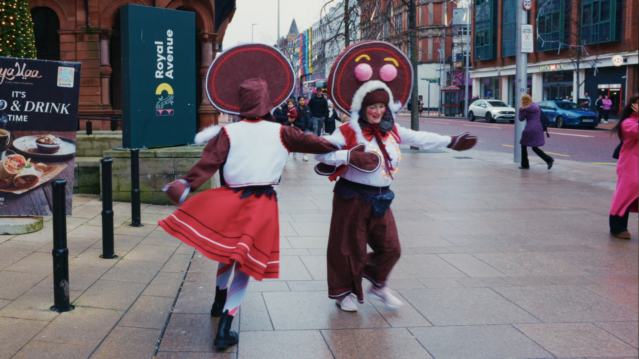 Two people dressed in festive costumes, including large gingerbread man and hats, dancing on a sidewalk in a busy city street during winter.