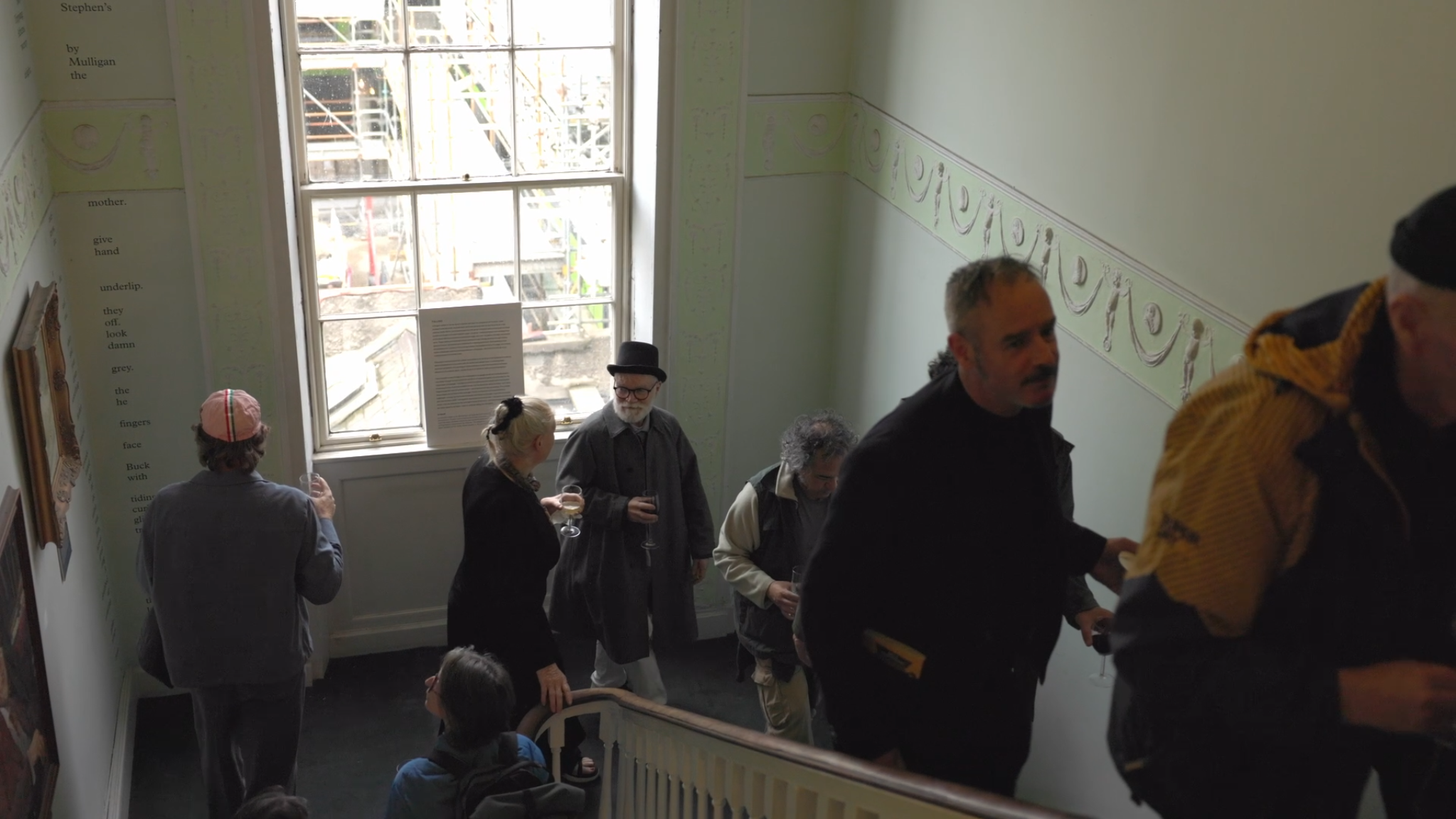 People attending an event inside the James Joyce centre, standing on a staircase, with a large window and decorative wall border.