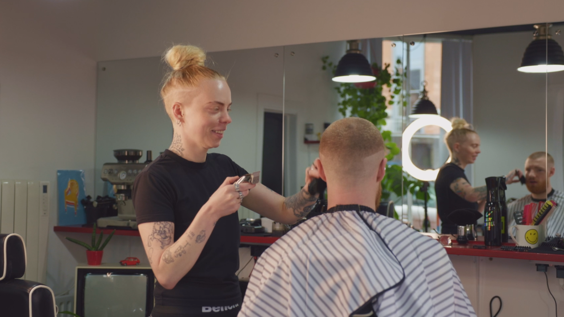 A hairstylist with blonde hair in a bun and tattoos is cutting a man's hair in a salon with a mirror reflecting the scene and a ring light in the background.