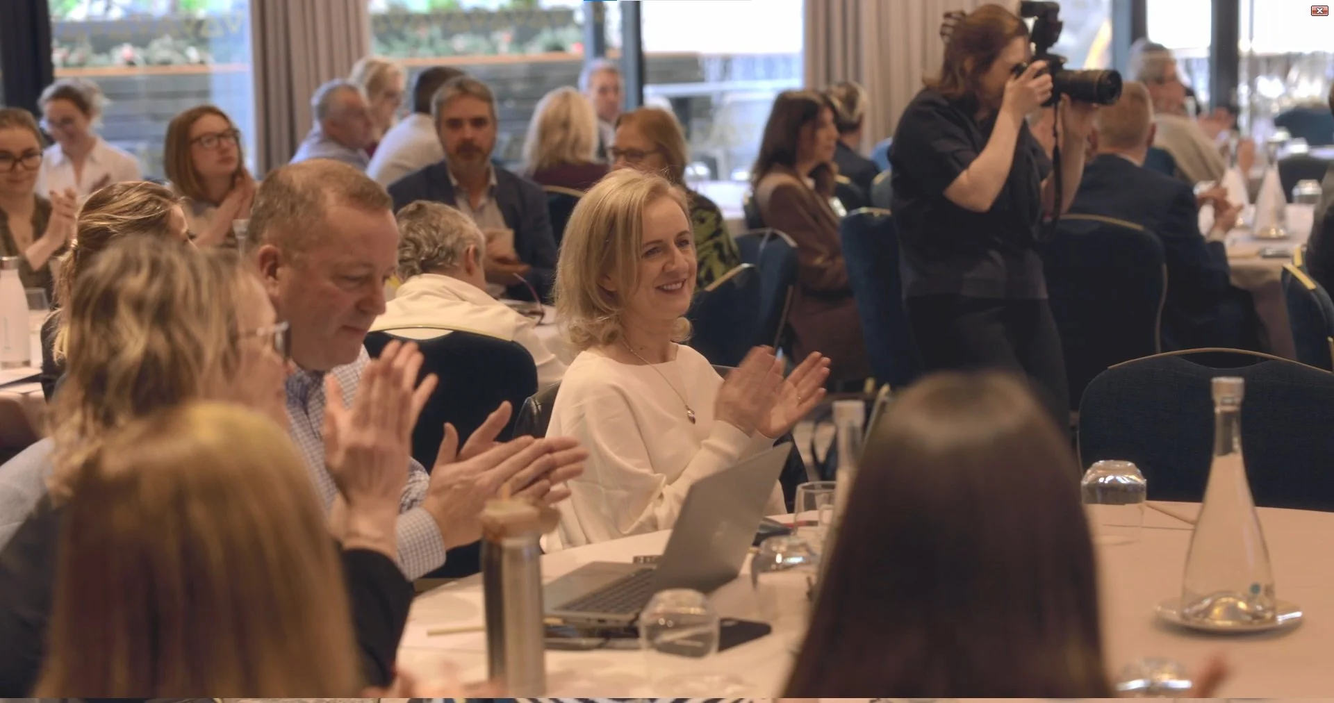 People sitting at round tables in a conference room, some clapping, a woman taking photographs, during an event or meeting.