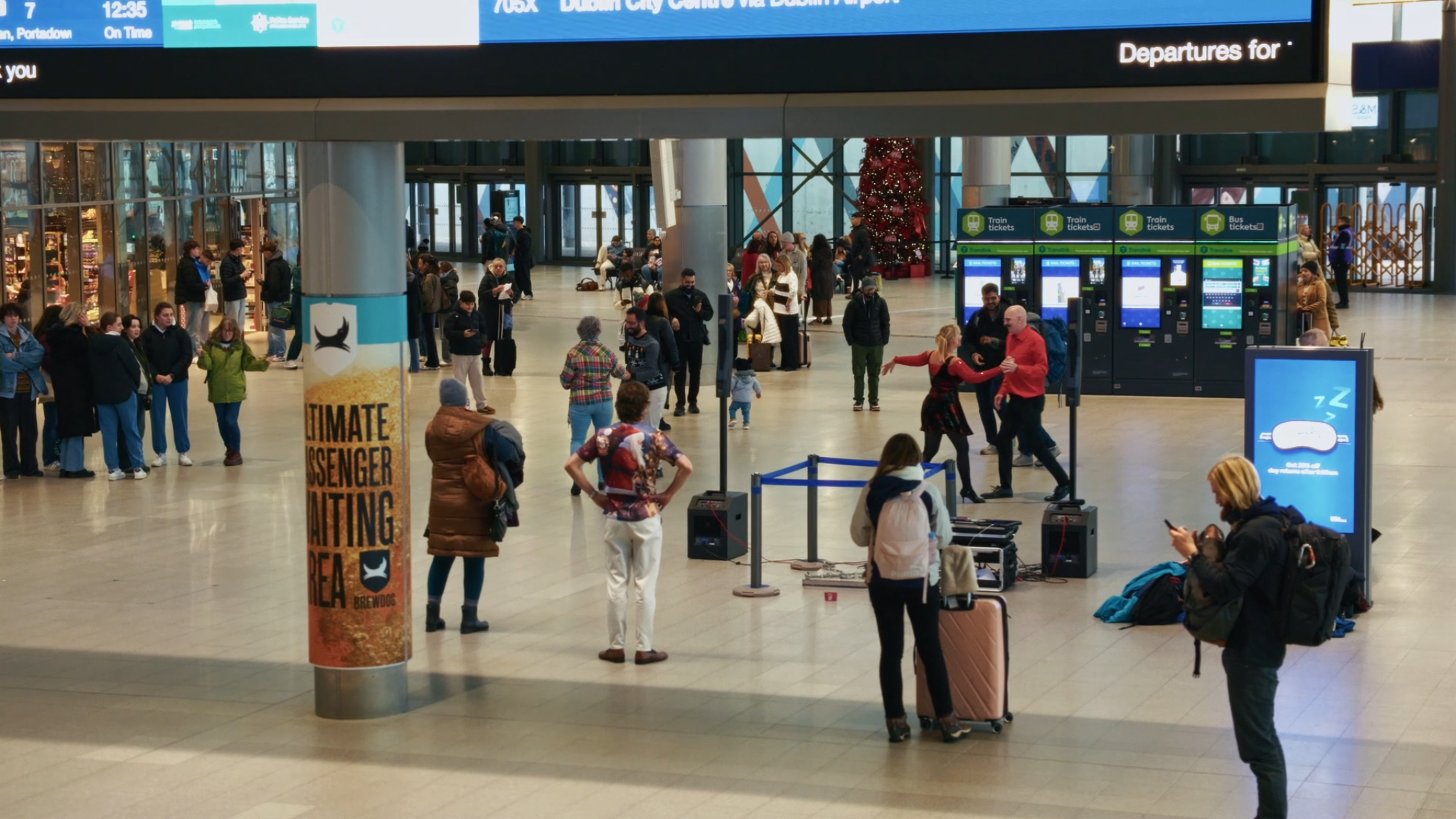 People waiting at a train station, some are standing in line, others are sitting or walking around. There are train ticket kiosks, a Christmas tree in the background, and a large digital flight information display overhead.
