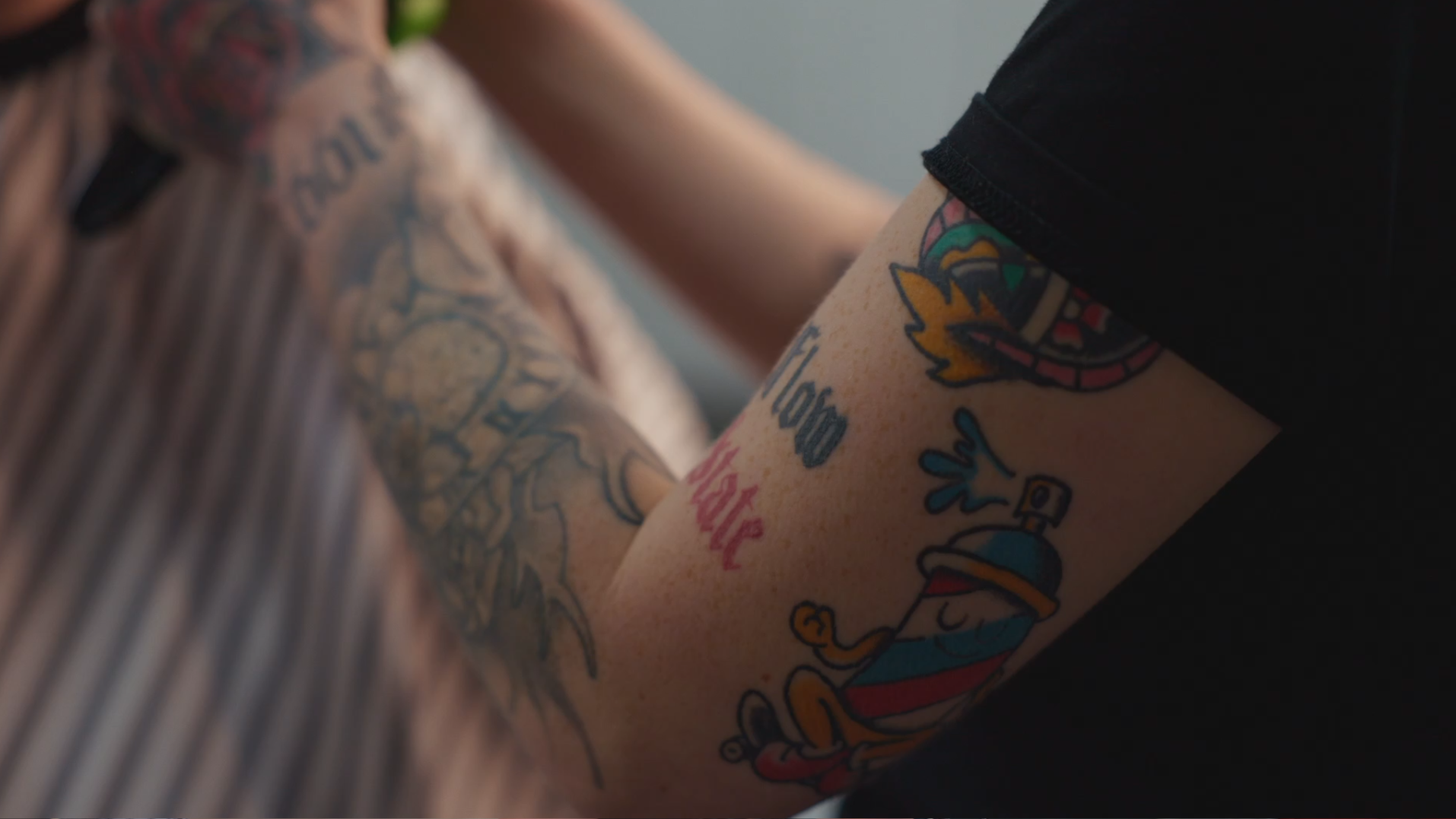 Close-up of arm tattoos, including a tattoo of a cartoon character with a spray bottle, and other colorful tattoo designs.