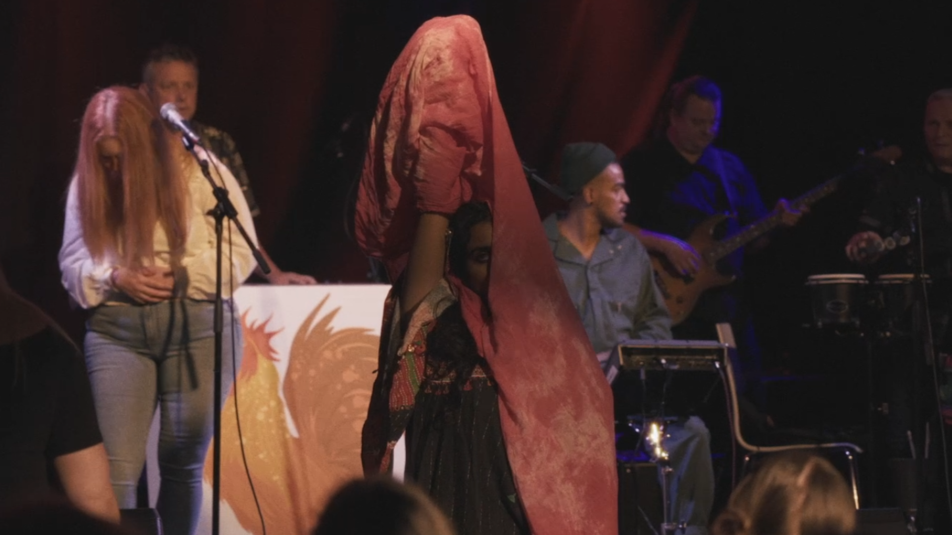 Person with face obscured by a large, red, textured fabric covering their head, standing in front of a musical band with guitars and drums, on a stage with a dark background.