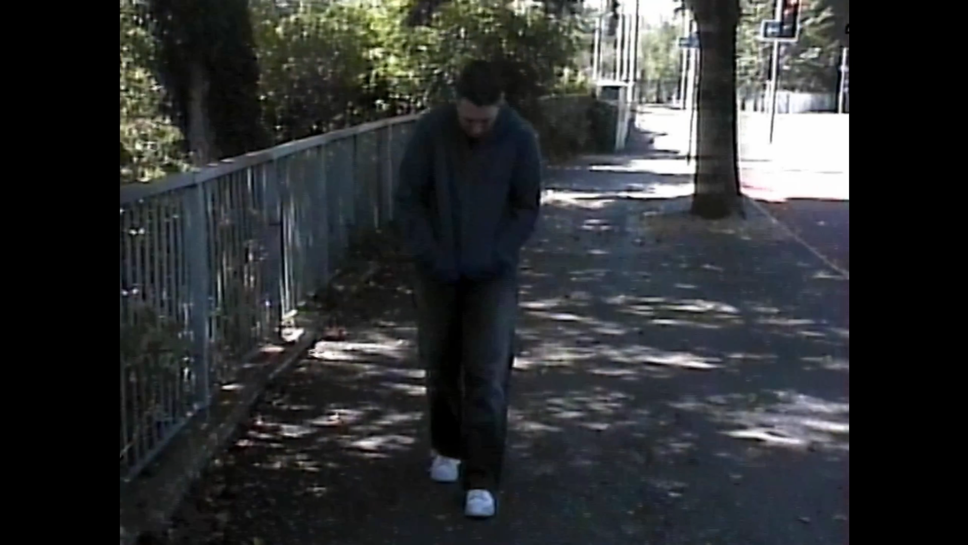 A young person walking alone on a sidewalk with trees and a metal fence on the side.