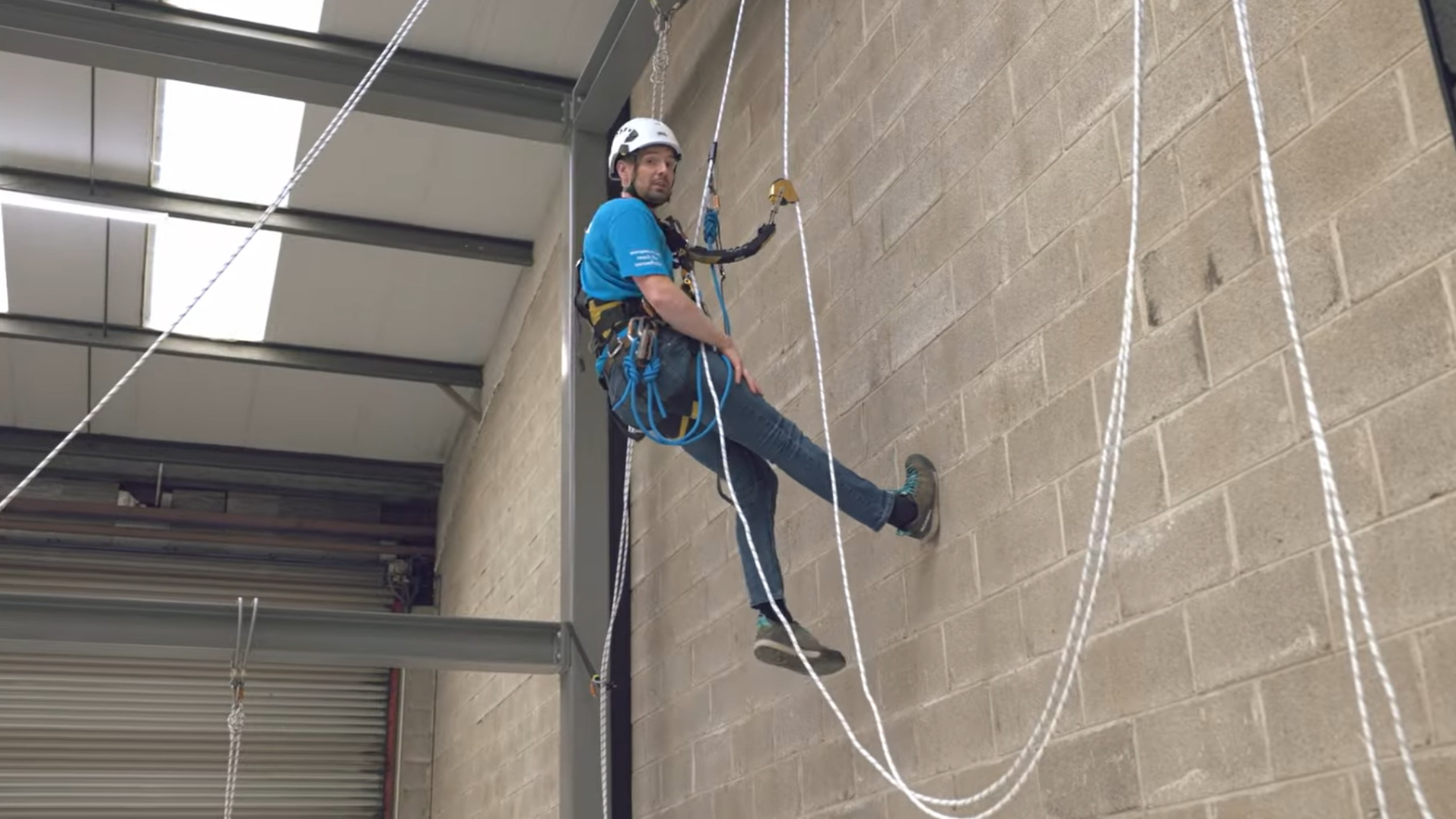A man in climbing gear and a helmet is practicing rappelling indoors on a wall with ropes.