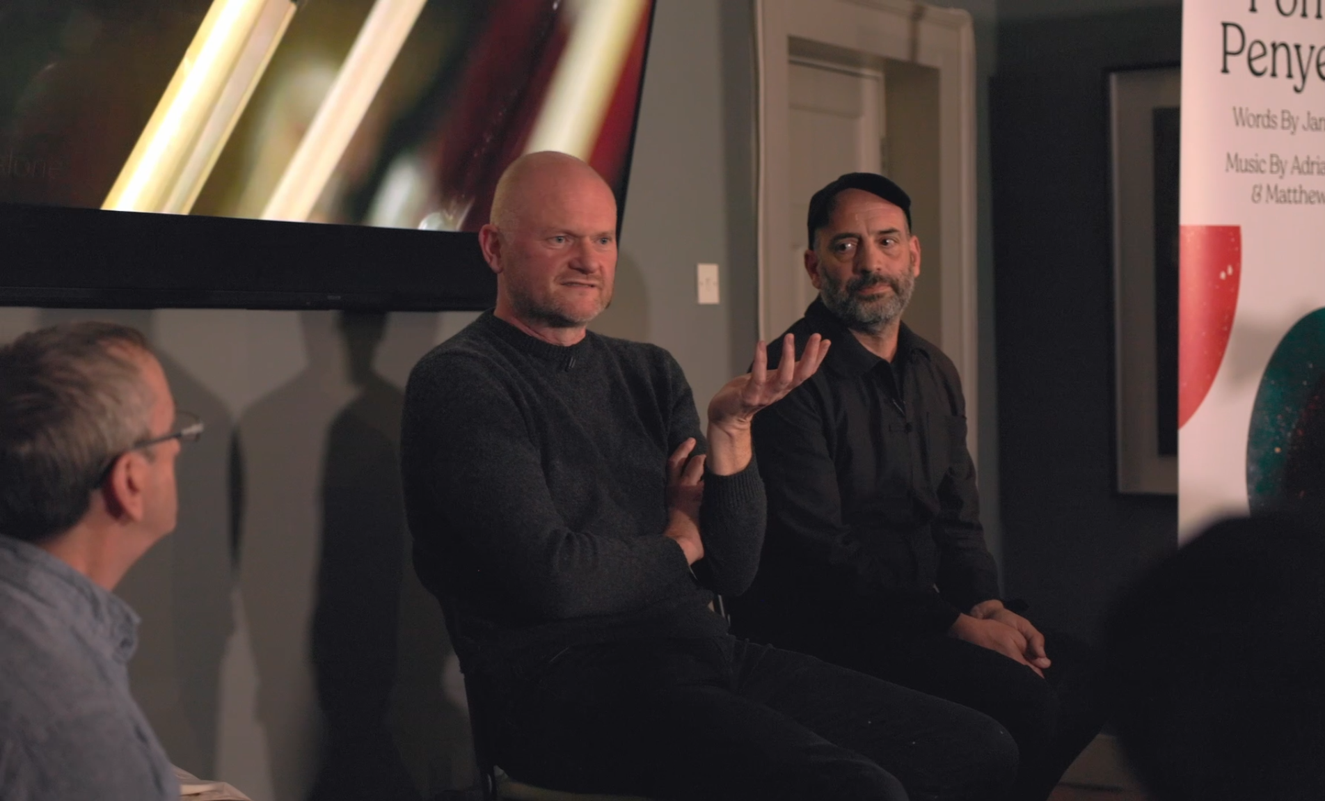 Three men sitting and talking at a panel discussion, with one man in the middle making a hand gesture and another man on the right listening, in a room with framed artwork and a colorful banner.