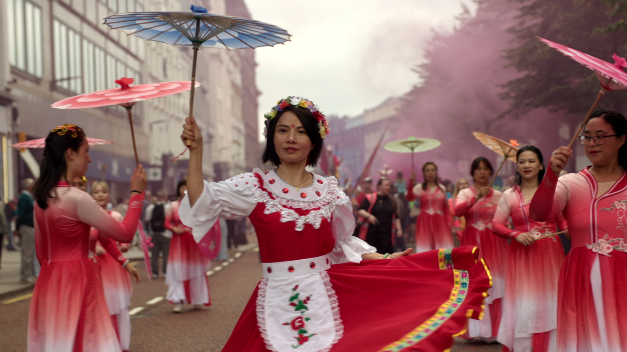 Women dressed in traditional pink and red dresses participating in a parade, holding umbrellas, with pink smoke in the background.