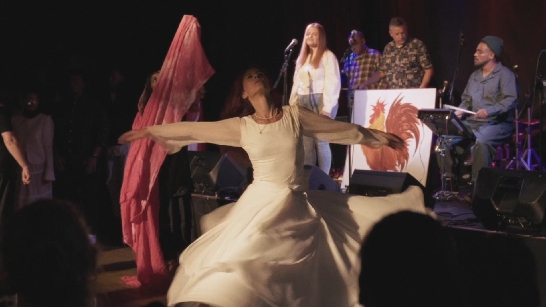 A woman in a white dress performing a dance on stage with her arms outstretched, while a singer stands behind her and a band plays, with a rooster illustration on the drum set.