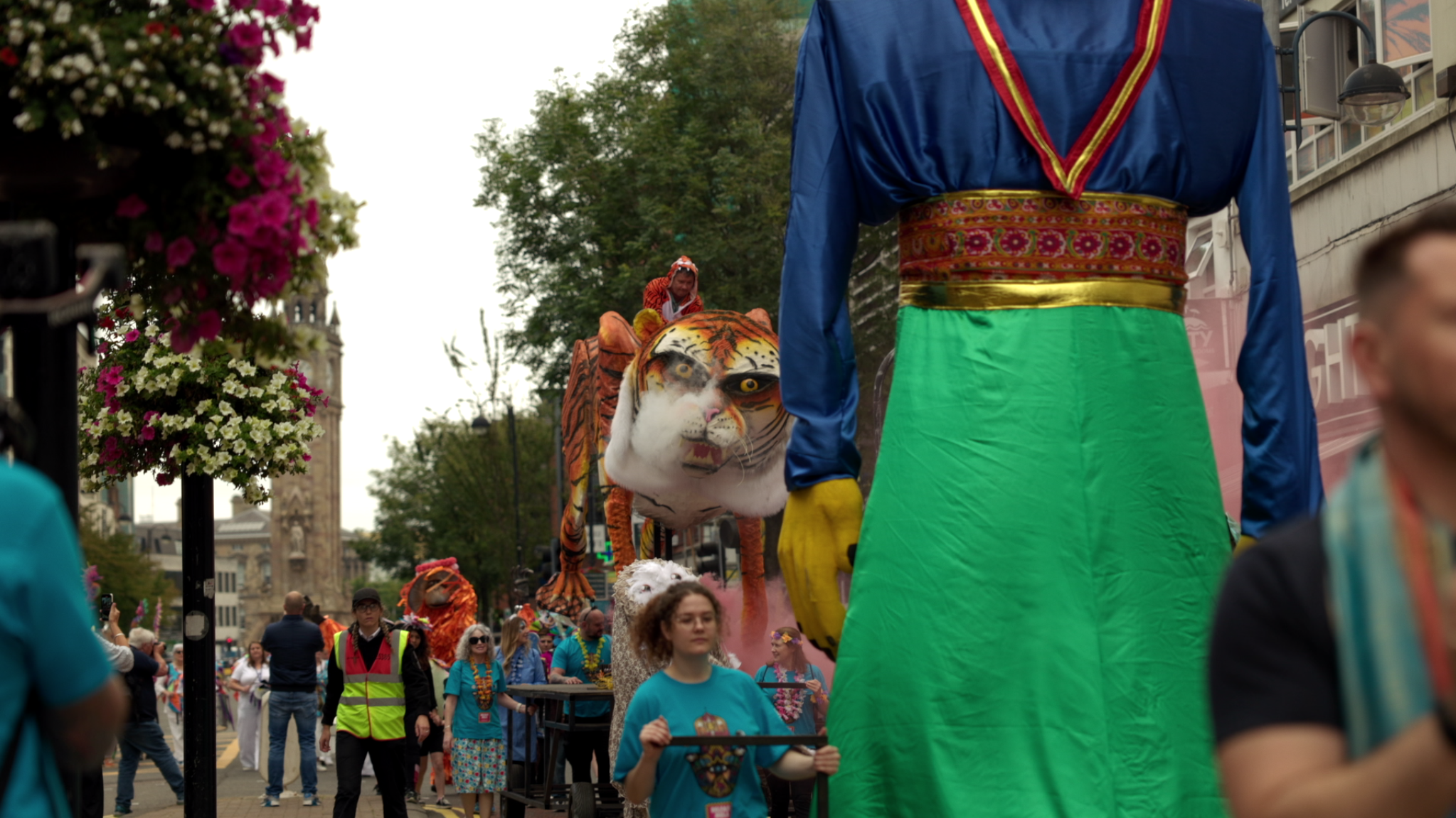 People participating in a parade with large colorful floats, including a tiger head and traditional costumes, on a city street lined with flowers and trees.