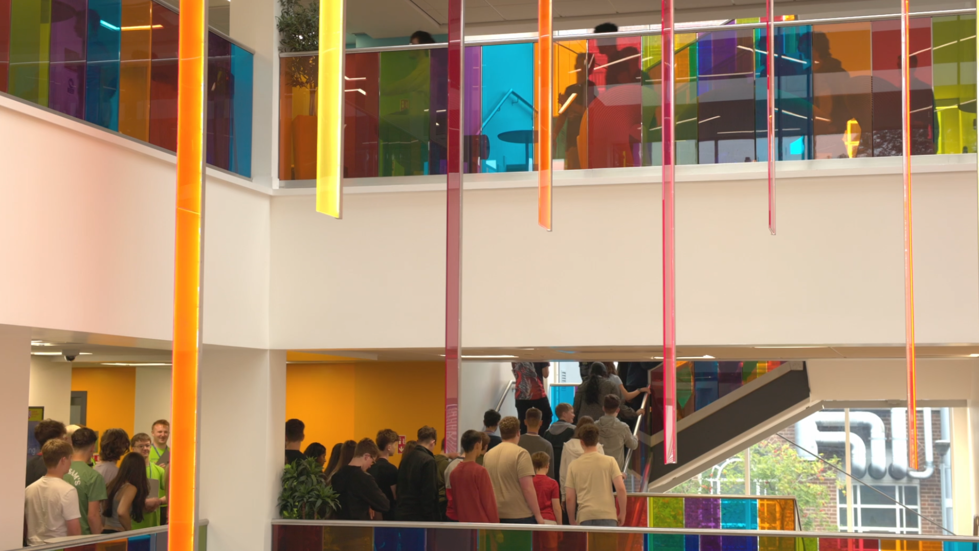 Group of people, mostly young adults, standing in line inside a colorful, modern building with bright rainbow-colored glass panels and decorative hanging strips.