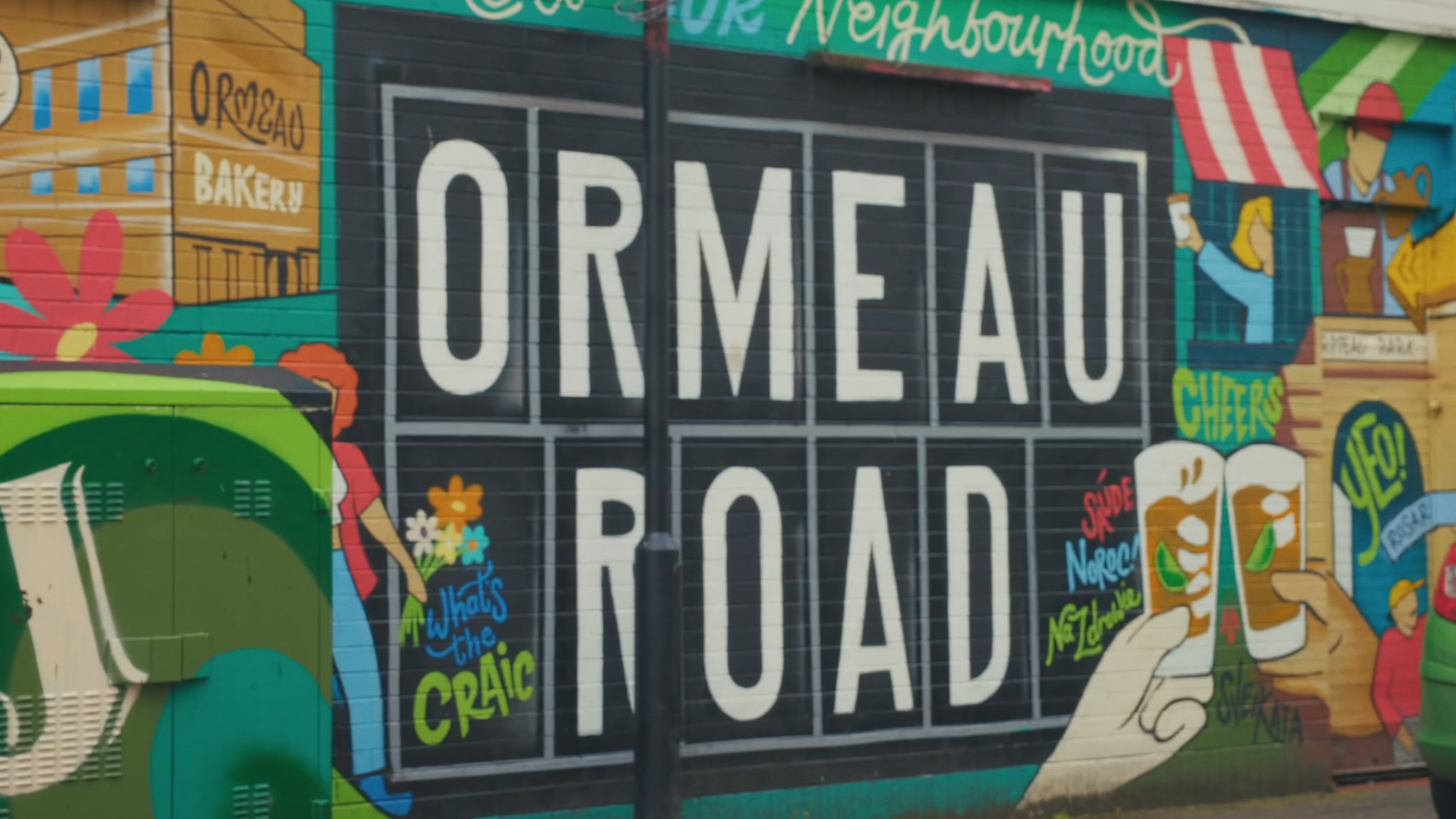 Colorful mural featuring the words 'Ormeau Road' and 'Neighbourhood' with illustrations of people, flowers, a green bus, and food items like beer glasses, along with phrases such as 'Cheers!', 'That's the Craic', and 'Skate North'.