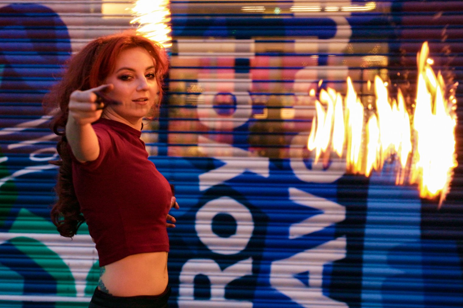 A young woman with long red hair pointing at the camera. She is wearing a burgundy top and standing in front of a graffiti-covered shutter with paint colors including green, blue, white, and black. There are flames on the right side of the image, add
