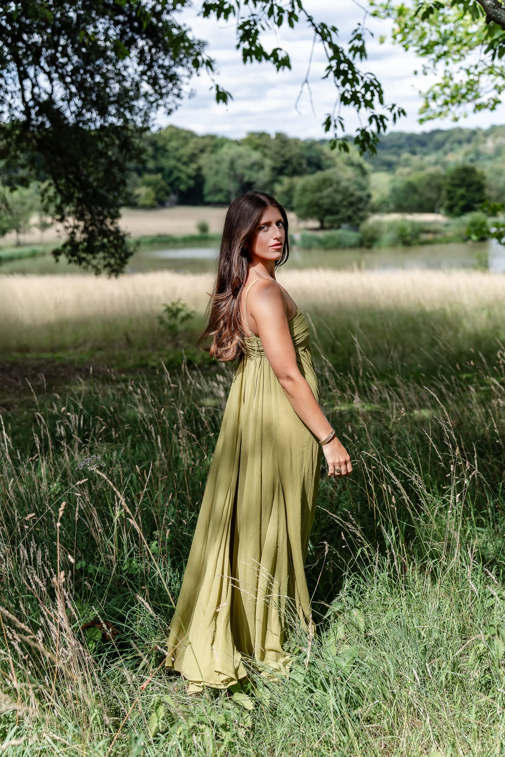 A woman in a flowing green dress standing in a grassy field near a river, with trees and a cloudy sky in the background.