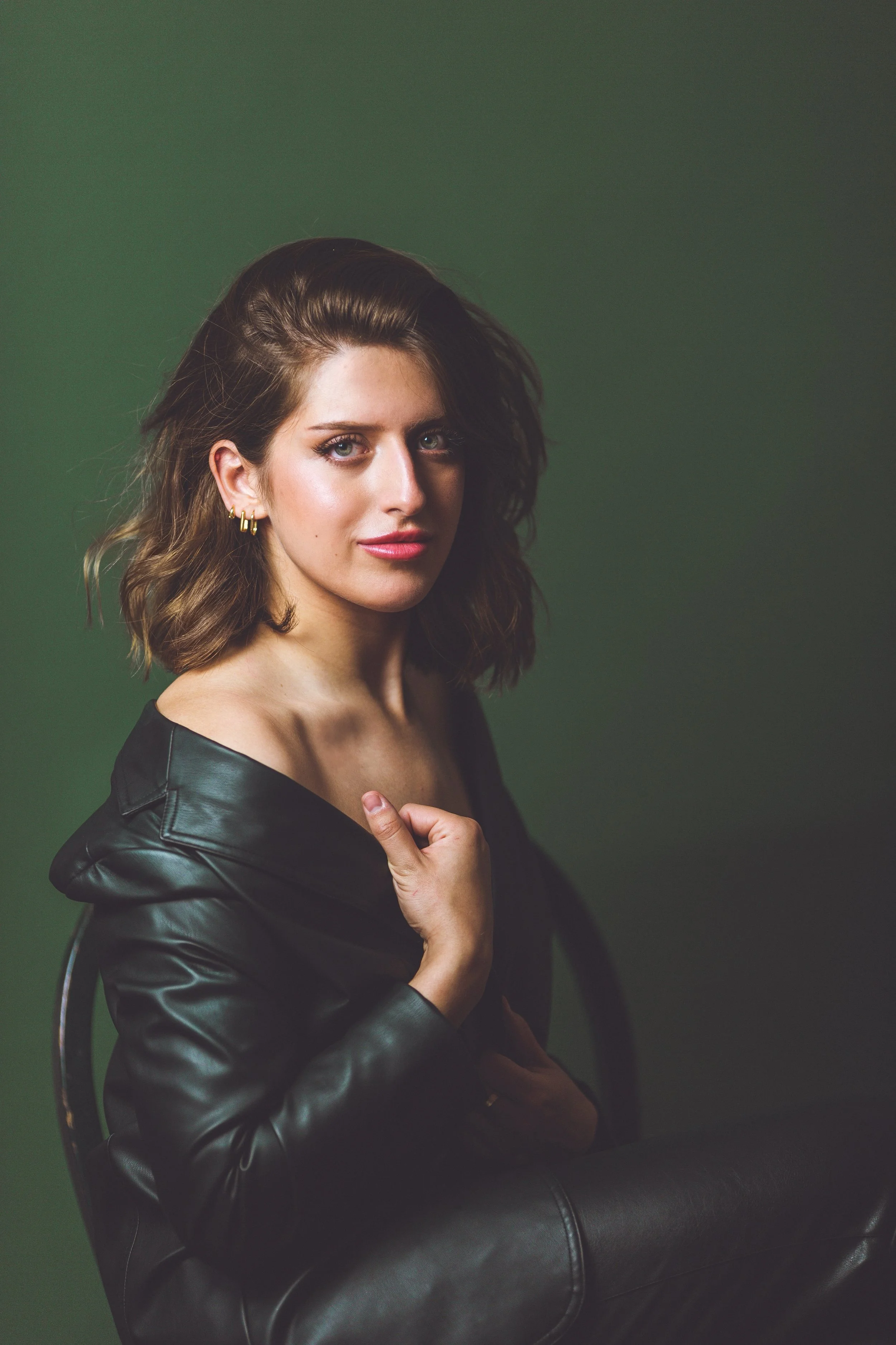 Portrait of a woman with shoulder-length brown hair, wearing a black leather jacket and earrings, posing against a green background.