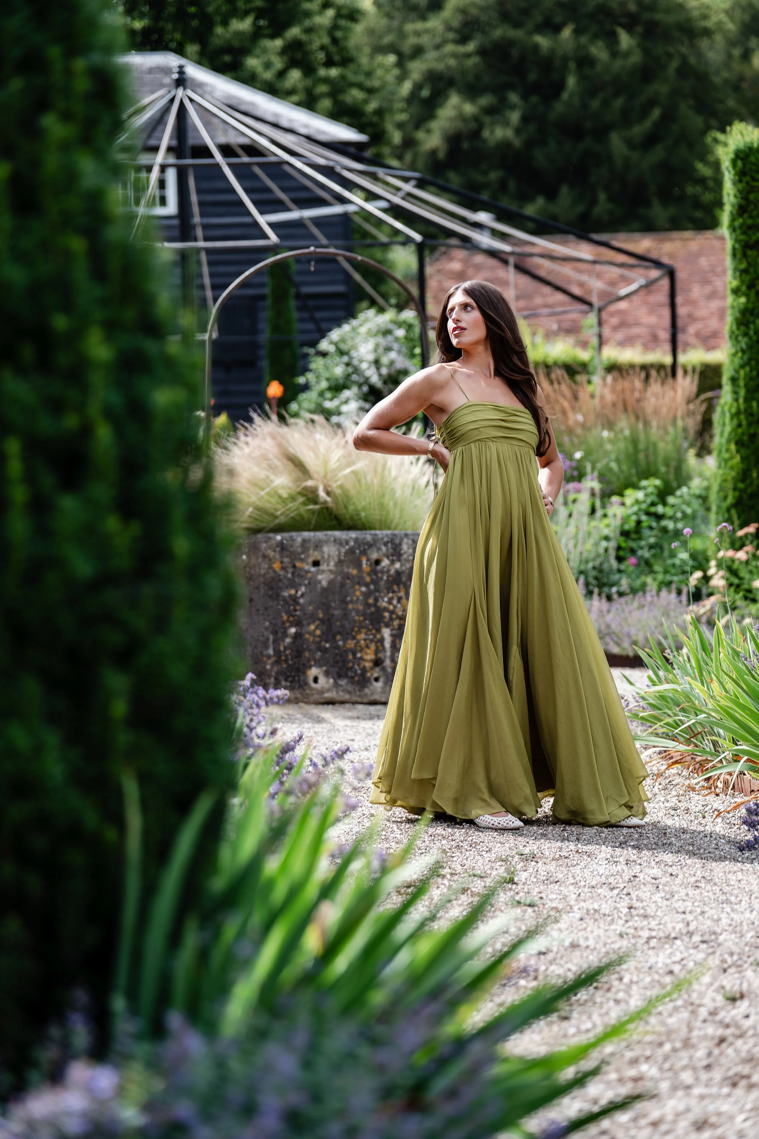 A woman in a flowing green dress stands in a lush garden with various plants and flowers, with a greenhouse structure and a dark wooden building in the background.