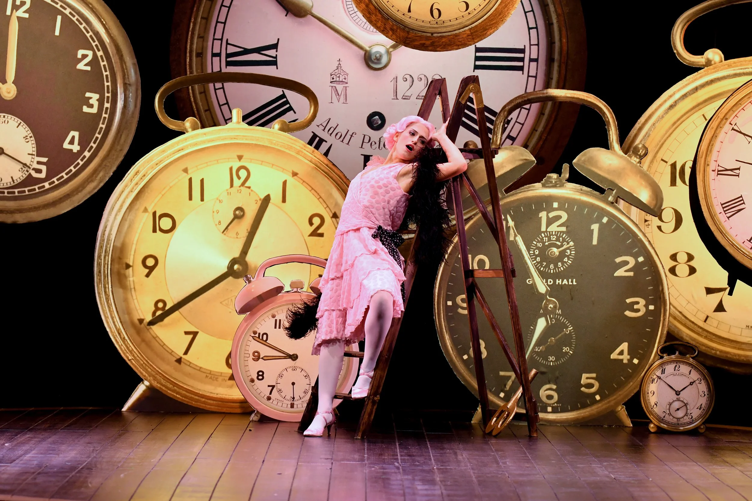 A woman in a pink dress and white stockings posing on a wooden ladder, surrounded by large vintage clocks of various sizes and colors