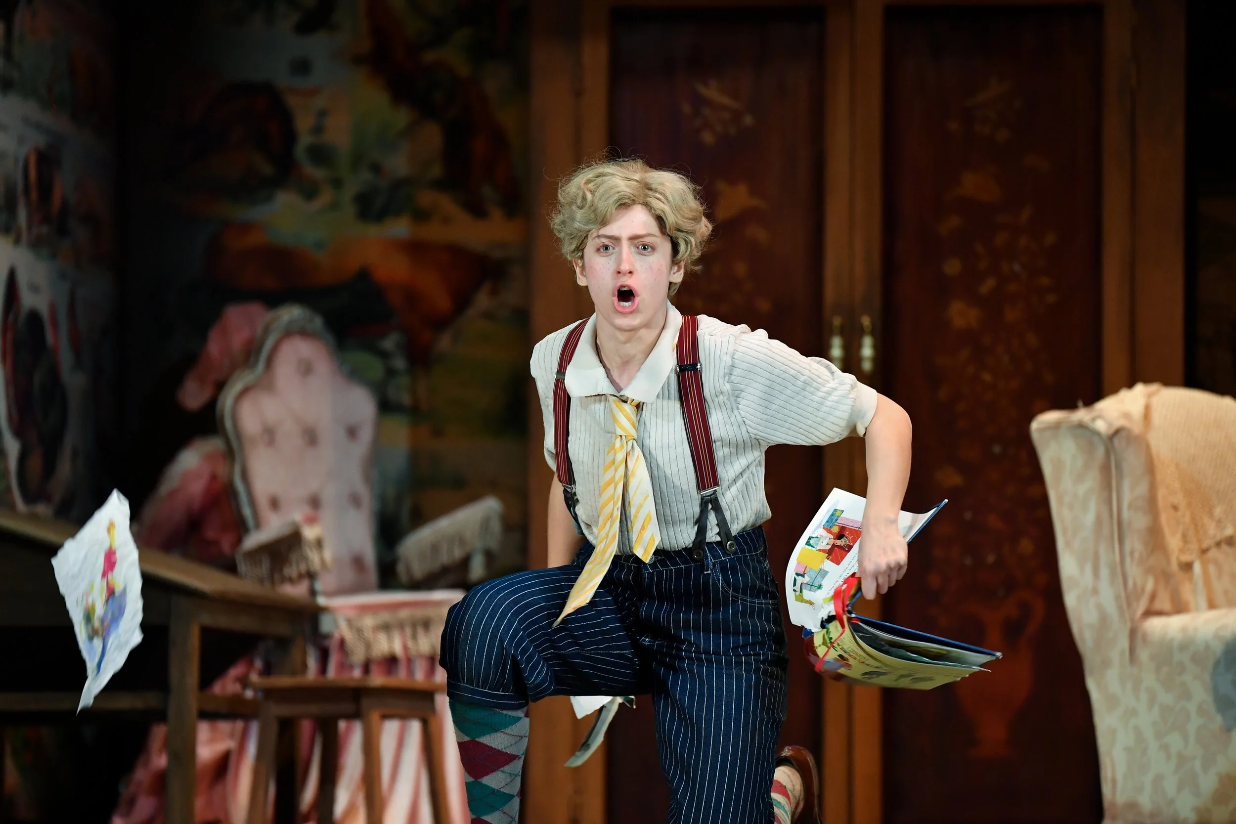 A young person with curly blonde hair, dressed in a striped shirt, suspenders, and pinstripe pants, shows an angry or upset expression while holding papers and running in a room with colorful artwork and vintage furniture.